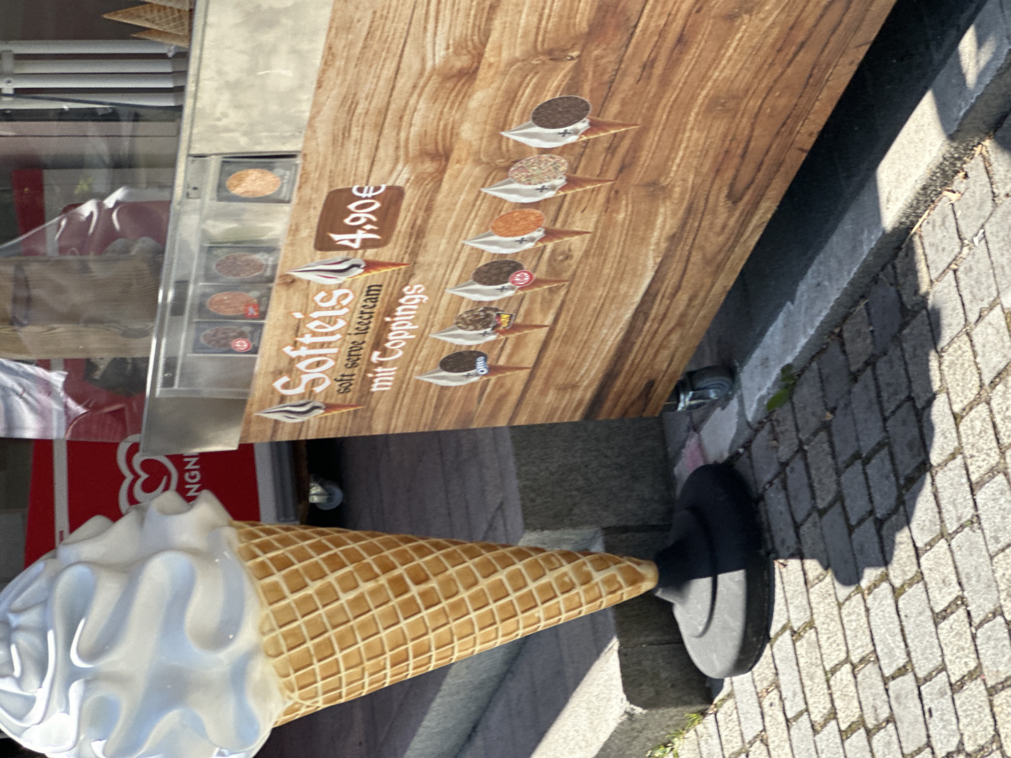 The image depicts an outdoor scene in Schönau am Königssee, Germany, featuring a soft-serve ice cream stand. A large, white ice cream cone statue stands to the left, partially obscuring a red and white vending machine with the "Langnese" logo. To the right, a wooden-grained sign advertises "Softeis" (soft serve ice cream) with the price of "4,90€". Below the price, there are illustrations of various soft-serve ice cream cones with different toppings, including what appears to be an Oreo cookie, a Kit Kat, and sprinkles. The stand itself has a metallic exterior and is positioned on a cobblestone pathway. The lighting suggests it is daytime with sunlight casting shadows. There are no people visible in the image.