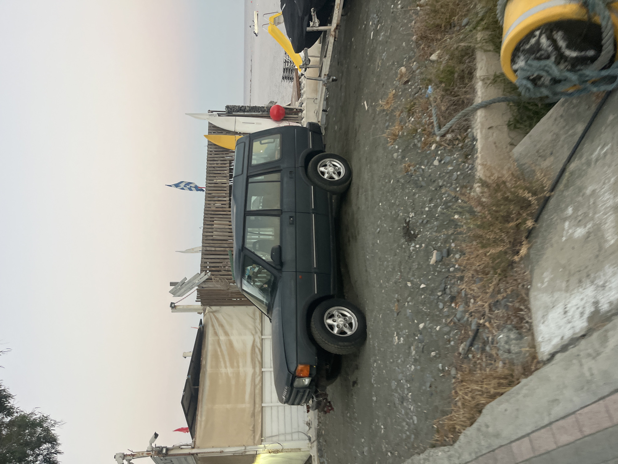 A dark green Land Rover Discovery is parked on a gravel lot near the beach in Agios Tychona Community, Cyprus. The vehicle is facing left and is partially obscured by a white tent-like structure on its left. Behind the car, there is a wooden fence, several surfboards or sailboards, and a red buoy. In the background, the sea is visible with a small white boat in the distance. The sky is a soft gradient of pale orange and white, suggesting it is either dawn or dusk. To the right of the Land Rover, a yellow slide leads into the water, and a boat covered with a black tarp is on a trailer. The foreground shows a concrete walkway with dry grass and pebbles.