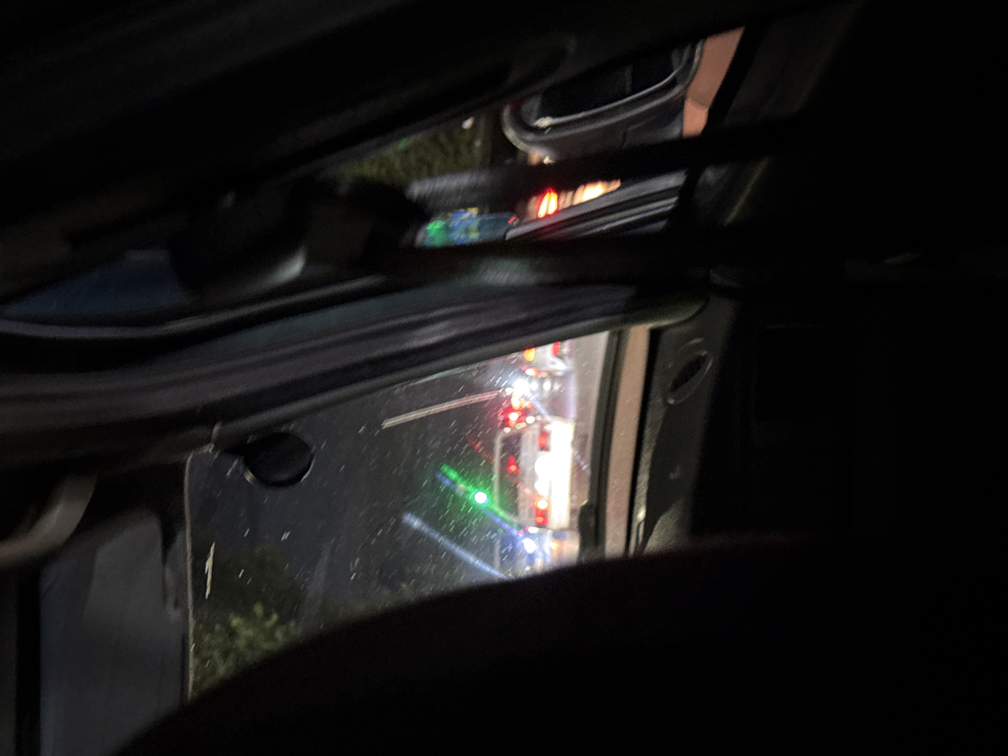 The image appears to be taken from inside a vehicle at night, looking out through a window. The primary subjects are a line of vehicles visible in the distance, indicated by their red taillights. There are also some colored lights, including green and blue, which might be from other vehicles or signage. The scene is dark, suggesting it's nighttime. The weather is unclear, but there are some reflections and streaks on the window that could indicate light rain or condensation.
The activity taking place is likely a traffic scene, possibly a traffic jam or a queue at a checkpoint or service area, given the stationary nature of the lights and the arrangement of vehicles. The perspective suggests the viewer is a passenger in one of the vehicles, observing the scene outside.
Notable details include the reflective surface of the window, which shows streaks and smudges. The surrounding environment outside the vehicles is largely obscured by darkness, though glimpses of greenery are visible, hinting at an outdoor, possibly roadside location. There is no visible text in the image. The overall mood is somewhat obscured and indistinct due to the low light conditions and the nature of the view through the window. Given the provided location context of Vladikavkaz, Russia, the scene is situated within this city, likely on a road or street.