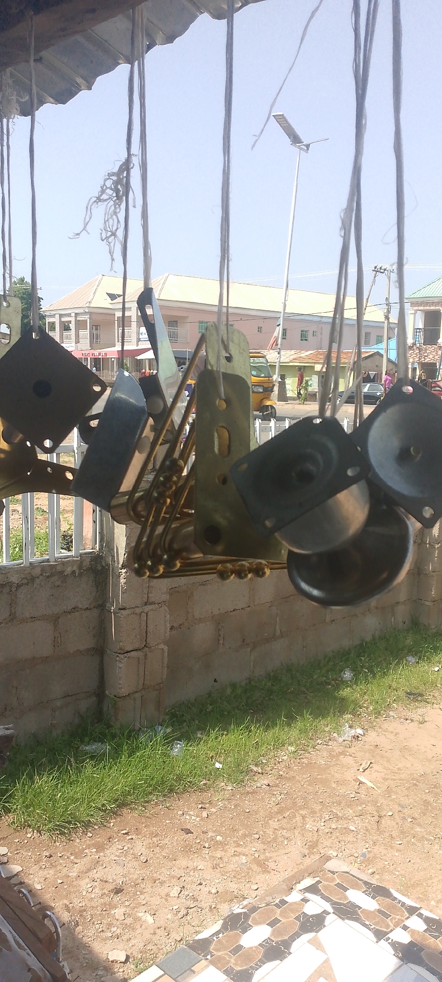 The image depicts a scene in Jalingo, Nigeria, likely a street or market area, on a clear day with a pale blue sky. The foreground is dominated by various metal objects, possibly spare parts or decorative elements, hanging from strings. These objects vary in shape and size, with some appearing to be flat plates with holes, and others being more rounded or cup-shaped. They are predominantly brass or a similar metallic material, with some showing signs of wear or patina.
In the middle ground, a low, light-colored brick wall runs horizontally across the frame, topped with a simple metal fence. Beyond the fence, green grass grows sparsely, with some scattered debris, including plastic bags. Further in the background, several buildings are visible. One prominent building is a two-story structure with a light yellow facade, balconies, and visible windows. To its left and right, other buildings are present, including a smaller green building. A yellow auto-rickshaw (a Keke Napep) is parked or moving on the road. A few people can be seen in the distance, walking near the buildings and the vehicle. A solar-powered street light is visible in the sky, mounted on a tall pole. The ground in the foreground is a mixture of dry soil, small stones, and a section of patterned tiling, suggesting it might be a pathway or part of a shop entrance. The overall impression is one of an outdoor commercial or workshop setting in an urban Nigerian environment.