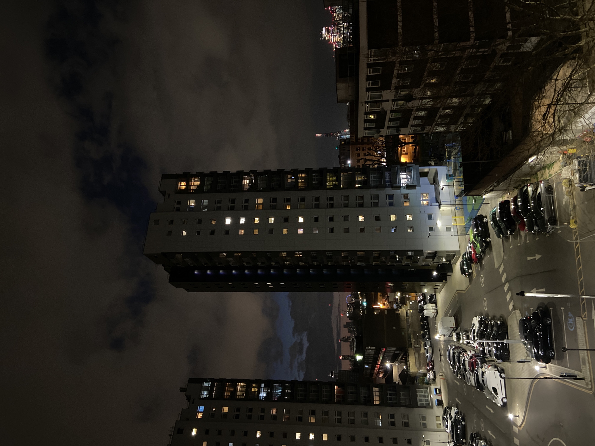 The image shows a nighttime view in London, United Kingdom, featuring tower blocks and a parking lot. Two tall residential buildings dominate the foreground, lit by numerous windows. A parking lot with various parked vehicles sits adjacent to the buildings. Beyond the immediate foreground, a cityscape with distant lights suggests the London skyline. The sky is cloudy.