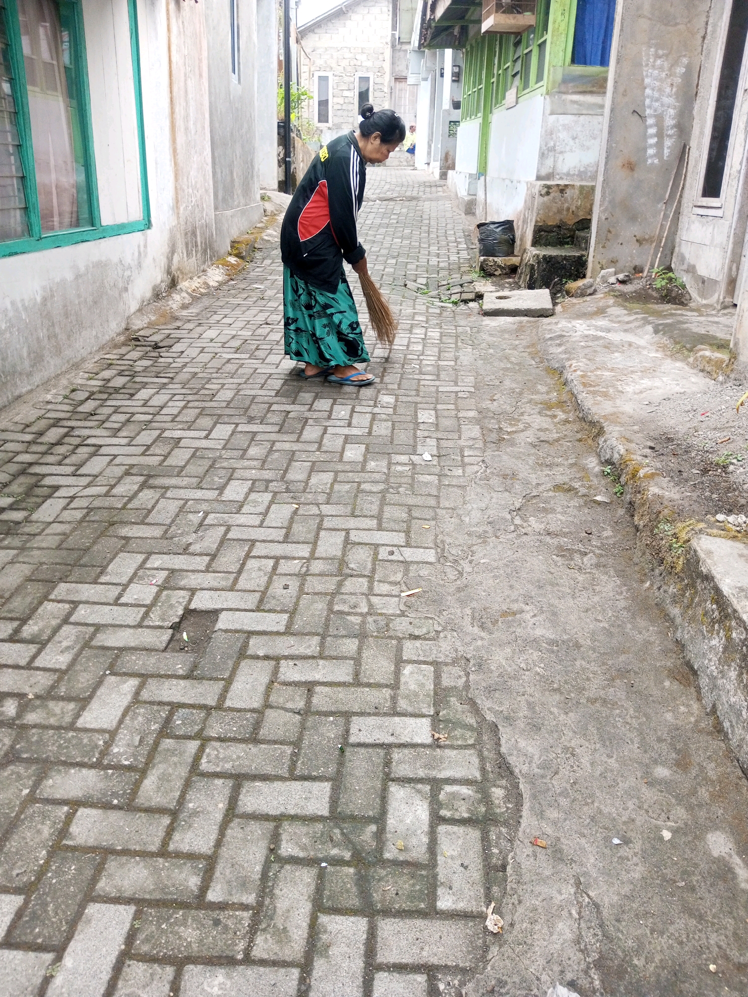 An Indonesian woman of middle age is sweeping a cobblestone alleyway in Andongsili, Indonesia. She wears a black and red athletic jacket and a long, green, patterned skirt. Her footwear consists of blue flip-flops. She is using a broom with a brown wooden handle and straw bristles. The alley is lined with buildings on both sides, constructed of concrete and brick. On the left, a building features a white facade with a large green-framed window. On the right, a building displays a rough, gray concrete exterior with several narrow, rectangular windows. The alleyway is paved with gray interlocking bricks, and a concrete path runs alongside it. Some small plants and debris are visible on the ground. The sky is not visible in the frame, suggesting an overcast or covered environment.
