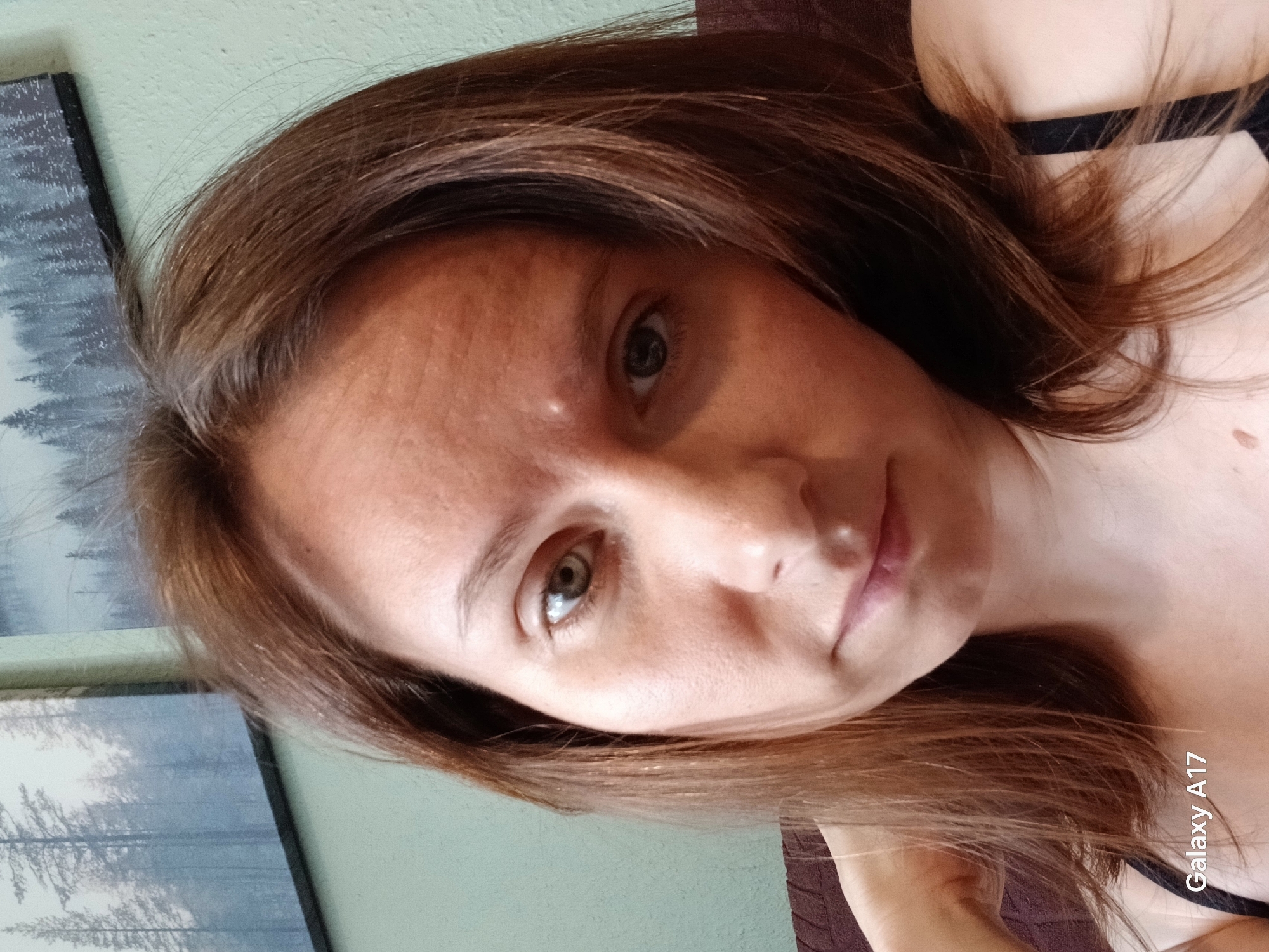 A close-up portrait of a woman with brown hair and light eyes, facing the camera. She appears indoors against a pale green wall with a framed picture on the left. The lighting is soft, and the photo has a slight downward angle. The camera used to take the photo is identified as Galaxy A17. There is no evidence to suggest this photo was taken in Yekaterinburg, Russia.