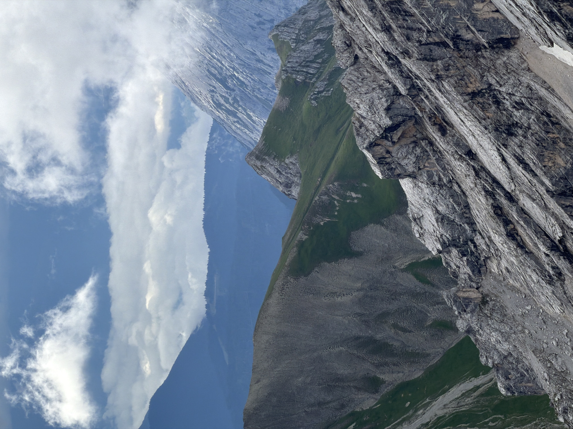 The image depicts a dramatic mountain landscape with multiple layers of peaks and ridges under a partially cloudy sky. The foreground is dominated by a steep, rocky mountain slope, characterized by weathered gray rock formations with distinct striations and smaller stones. To the left and middle ground, there are gentler slopes covered in green grass, interspersed with rocky outcrops. Further in the background, a large, mist-shrouded mountain peak rises, its upper reaches obscured by clouds. The sky above is a mix of bright blue and white clouds, some of which are wispy and others denser. The scene is devoid of any human presence or man-made structures, presenting a raw and natural alpine environment. This is likely a view of the Alps, possibly from a vantage point near Garmisch-Partenkirchen, Germany, given the characteristic terrain and vegetation.