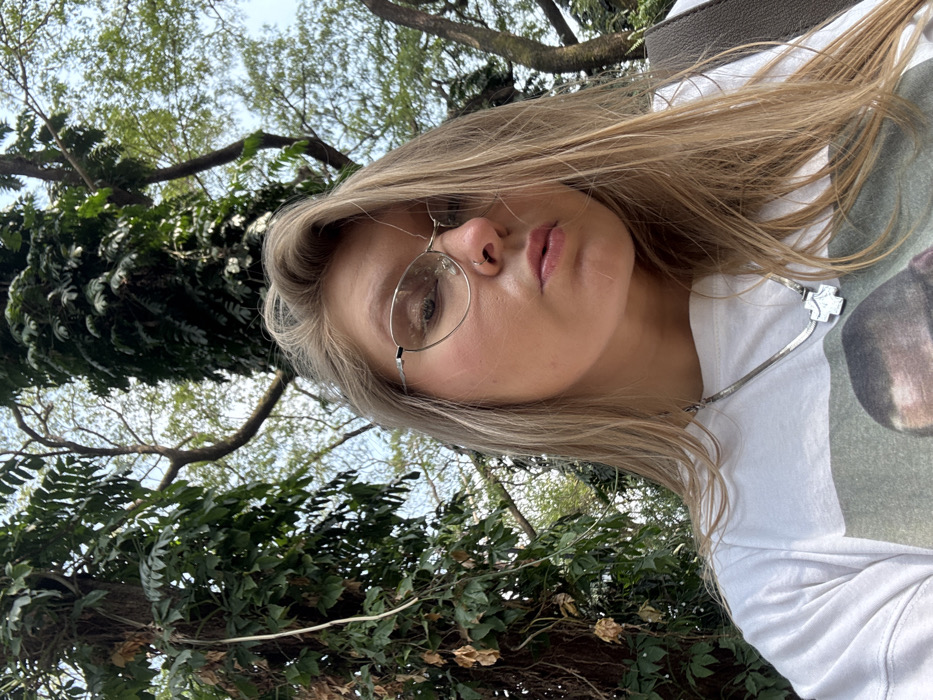 A person with blonde hair and round glasses gazes towards the camera. The individual wears a white t-shirt and a silver necklace with a cross-shaped pendant. Lush greenery, including tree branches and foliage, surrounds the subject. The background suggests an outdoor setting, possibly a park or garden. The photo's angle is slightly upward, capturing the person's face and upper body.