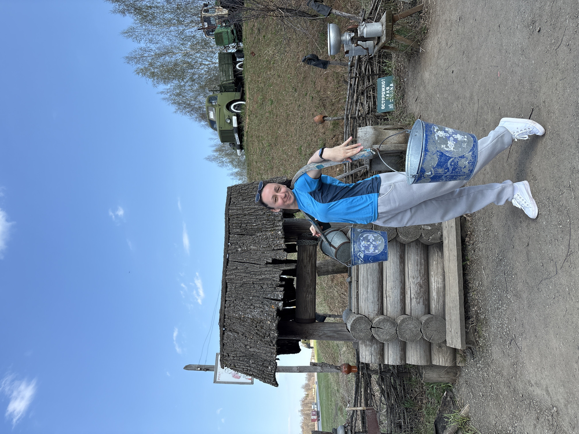 A woman is standing in front of a wooden well in Bol'shoi Mashlyak, Russia. She is wearing a blue shirt, gray pants, and white sneakers. She is holding two blue buckets. Behind her are several vintage military vehicles and a sign in Russian. The sky is blue with scattered clouds. The ground is dirt.