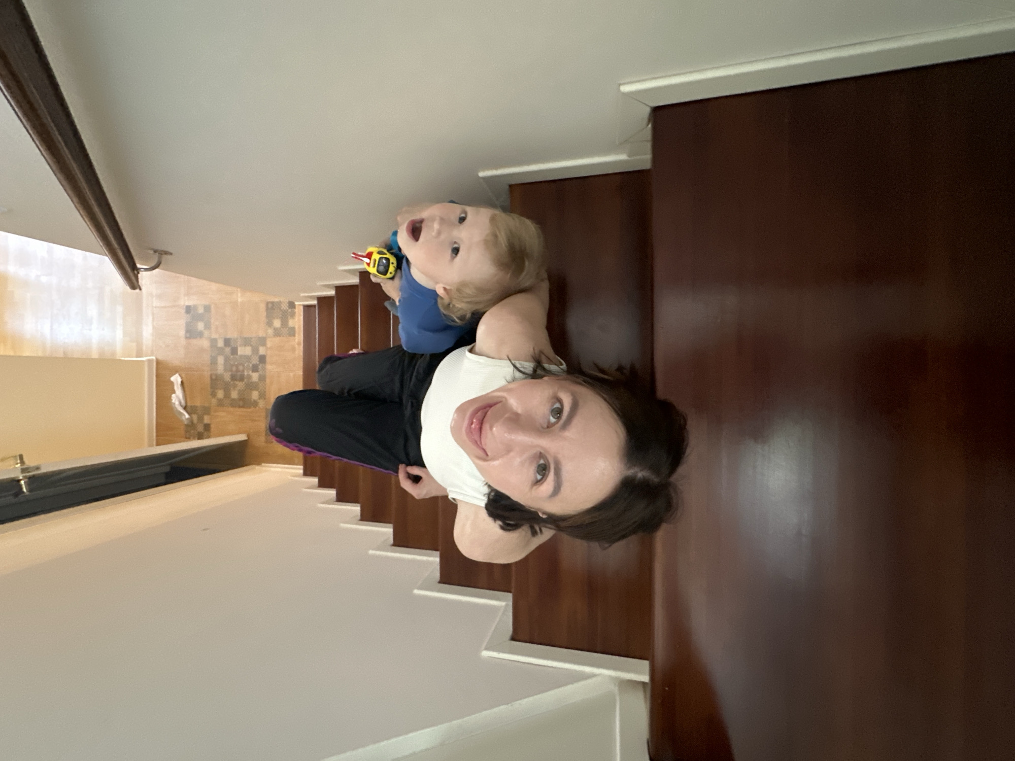 A woman and a young child are standing on a staircase in an indoor environment. The woman is wearing a white shirt and black pants, and she is looking directly at the camera with a smile. The child, who is being held by the woman, is wearing a blue shirt and holding a yellow and black toy. The staircase has dark brown wooden treads and white risers. The floor at the bottom of the staircase is covered in patterned tiles. The walls are white. The location appears to be a home interior. There is no specific information to confirm the city of Русский Вожой, Russia.