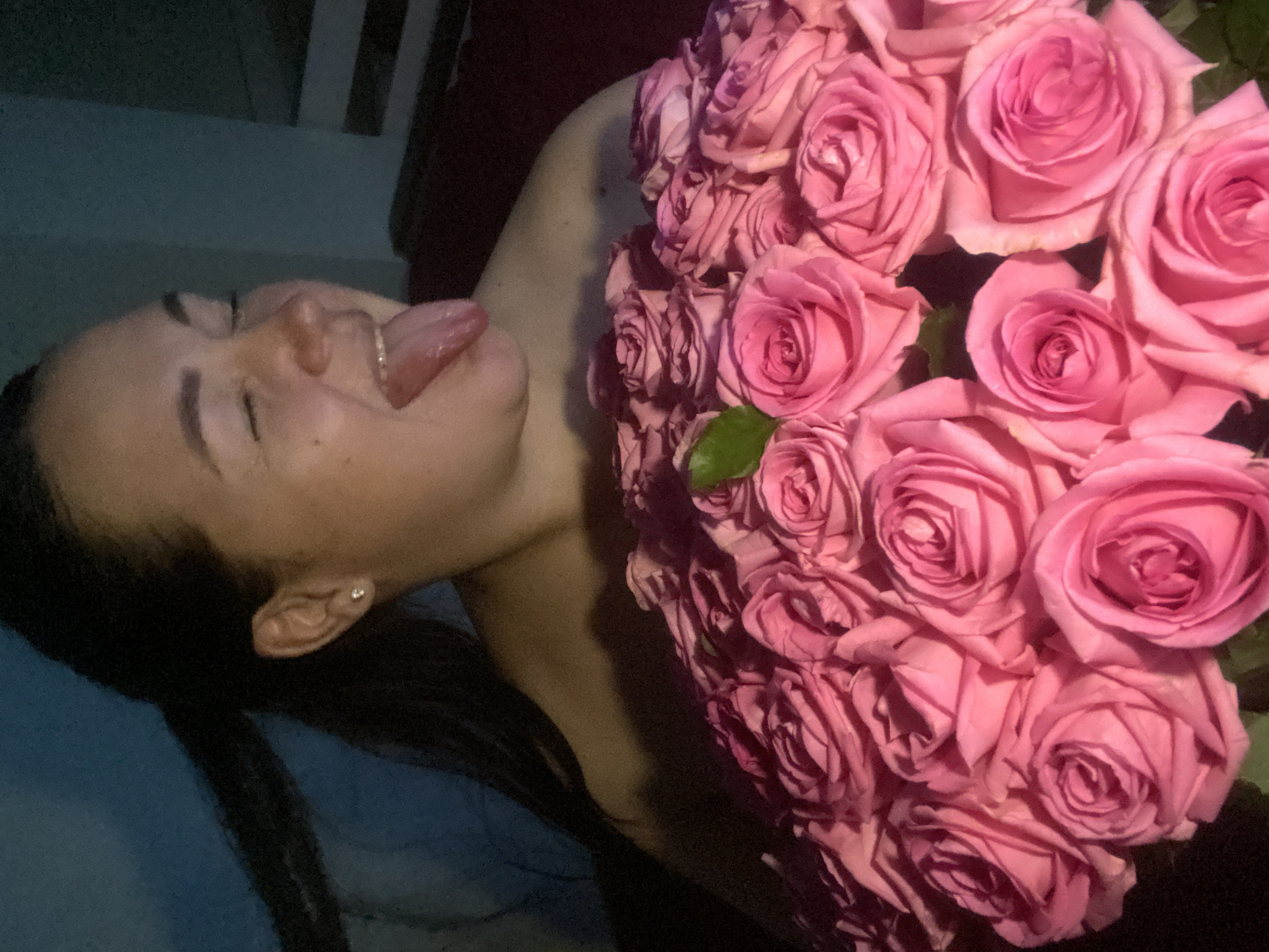 A woman with dark hair and fair skin is holding a large bouquet of pink roses. Her eyes are closed, and her tongue is sticking out. She is wearing a small earring in her left ear. The background is dark and indistinct. There is no evidence indicating this image was taken in Sochi, Russia.