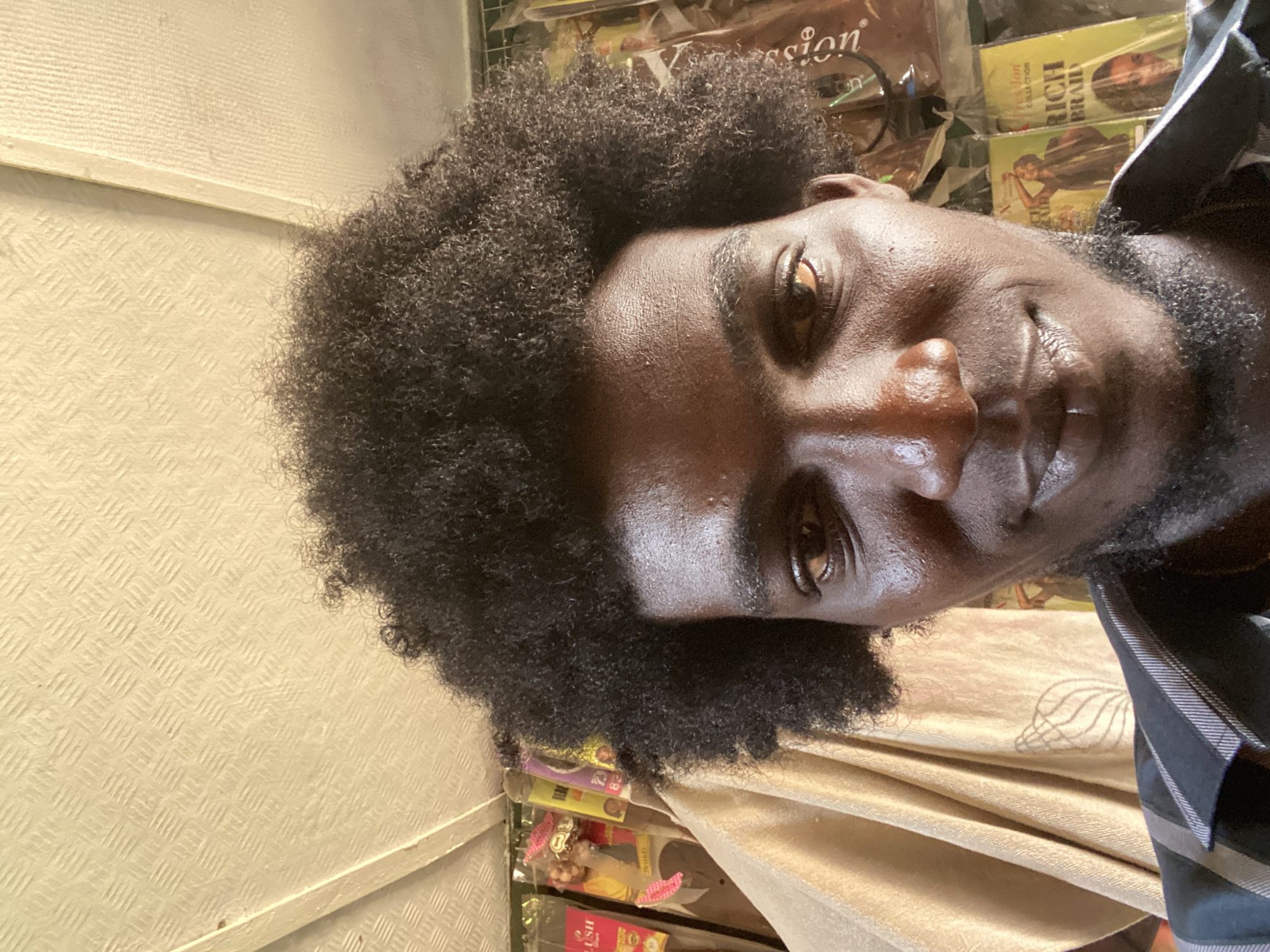 A man with dark skin and short, curly black hair is pictured. He appears to be in his mid-20s to early 30s. He is wearing a patterned collared shirt and is in what looks to be a retail environment, possibly a store selling hair products or beauty supplies. This is indicated by the stacks of hair extensions and braiding products visible in the background. The location is likely in Lagos, Nigeria, based on the products and general aesthetic. The photo has a casual, selfie-style feel.
