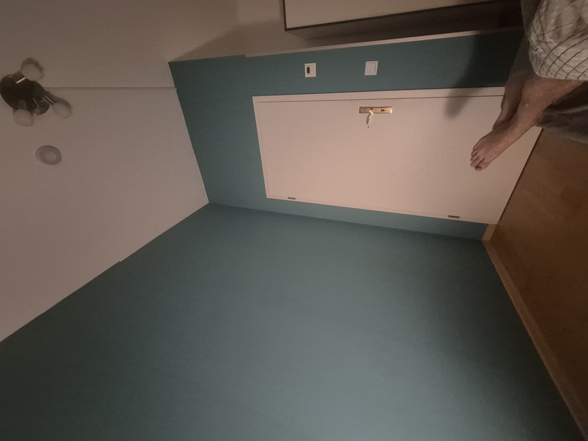 The image shows a corner of a room with teal walls and a wooden floor. A white door with a gold handle is visible on the right side of the frame. A person's legs and feet are in the foreground, resting on a blanket. The person's feet are crossed, and one foot has dark nail polish. Above the door, there are two white wall plates, one of which appears to be a light switch. A ceiling fan with multiple lights is visible in the upper right corner. A circular object, possibly a smoke detector, is on the ceiling to the left of the fan. The scene appears to be indoors, likely in a bedroom or living space. Based on the provided information, this location is Papendorf, Germany.