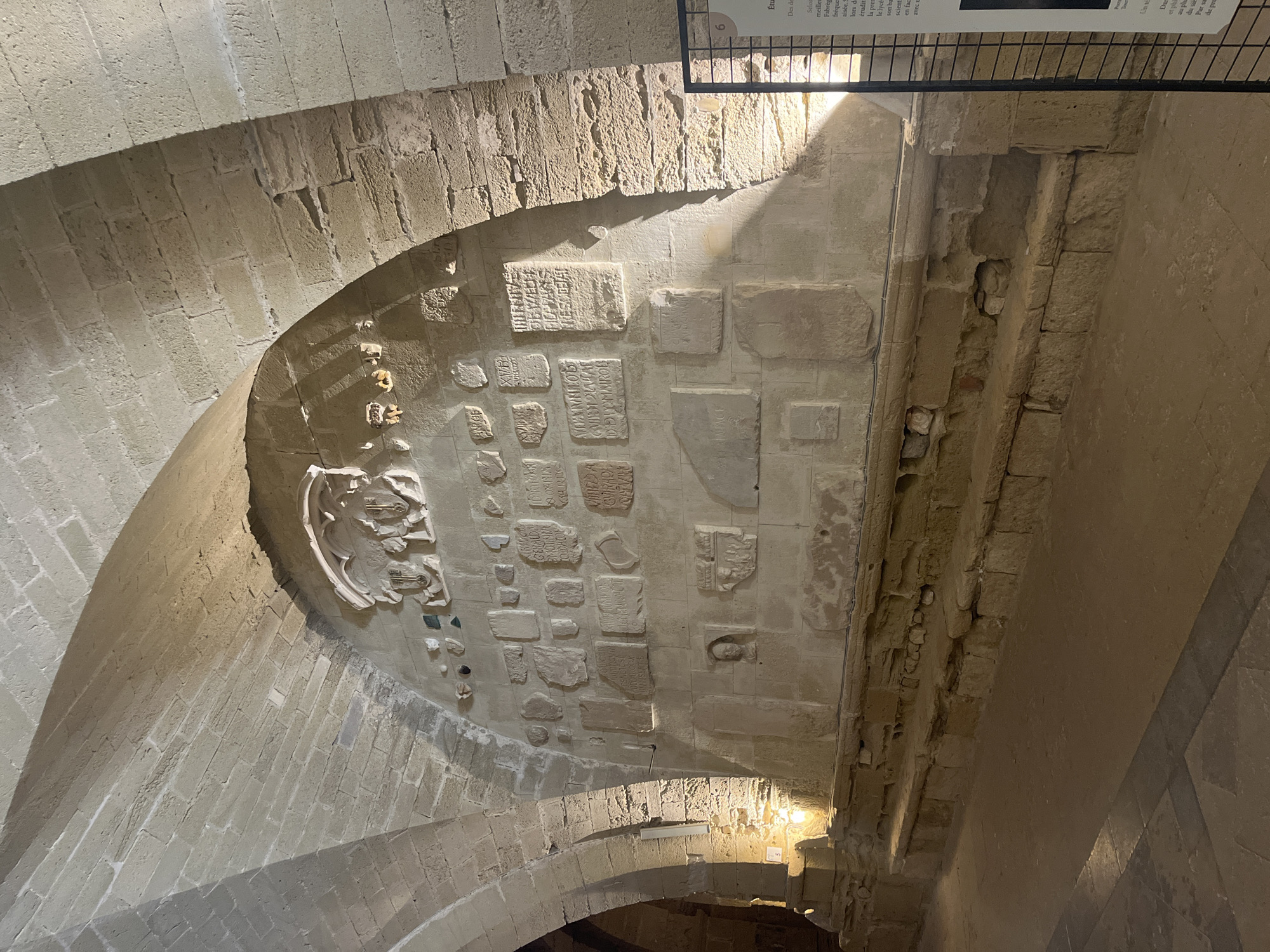 The image depicts an interior view of the Romanesque bell tower of the Cathédrale Saint-Jean-Baptiste de Maguelone, located in Villeneuve-lès-Maguelone, France. The scene is characterized by rough-hewn stone architecture, including arched ceilings and a stone wall adorned with numerous carved stone tablets and artifacts. The wall features a central escutcheon with three shields, and below it, a variety of rectangular and irregular stone slabs bearing inscriptions and decorative carvings. A bust or carved head is also visible embedded in the wall. The floor is paved with stone tiles. Stone benches or seating are present along the base of the wall. Lighting appears to be artificial, casting shadows and highlighting the textures of the stone. A metal exhibition panel with text and images is partially visible on the right side of the frame. The overall environment suggests an archaeological or historical exhibit within the bell tower.