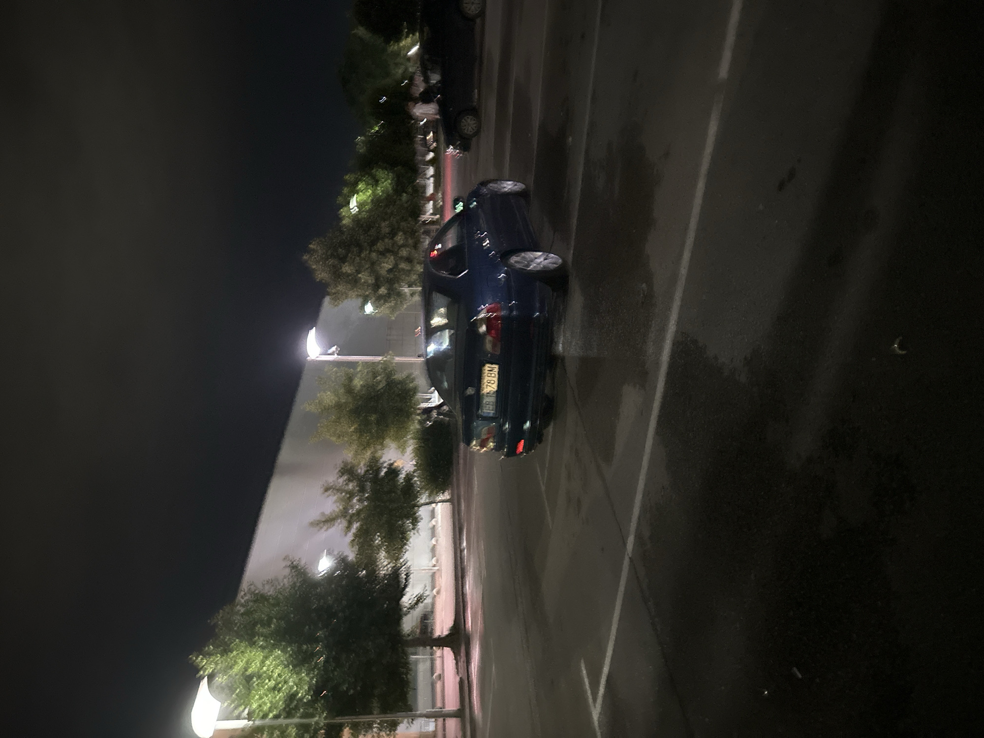 The image captures a dark, wet parking lot at night, possibly in the city of Ruse, Bulgaria, given the license plate "EB 7578 BM". A dark blue sedan, likely a BMW, is parked facing right, its rear visible. The car is illuminated by streetlights, which also cast reflections on the wet asphalt. Behind the sedan, another dark vehicle is partially visible, also parked. In the background, there are several trees and a long, light-colored building. The sky is dark and overcast, suggesting nighttime. A few indistinct figures appear to be walking in the distance near the building and trees. The scene conveys a sense of stillness and low visibility typical of a dimly lit urban environment after dark.