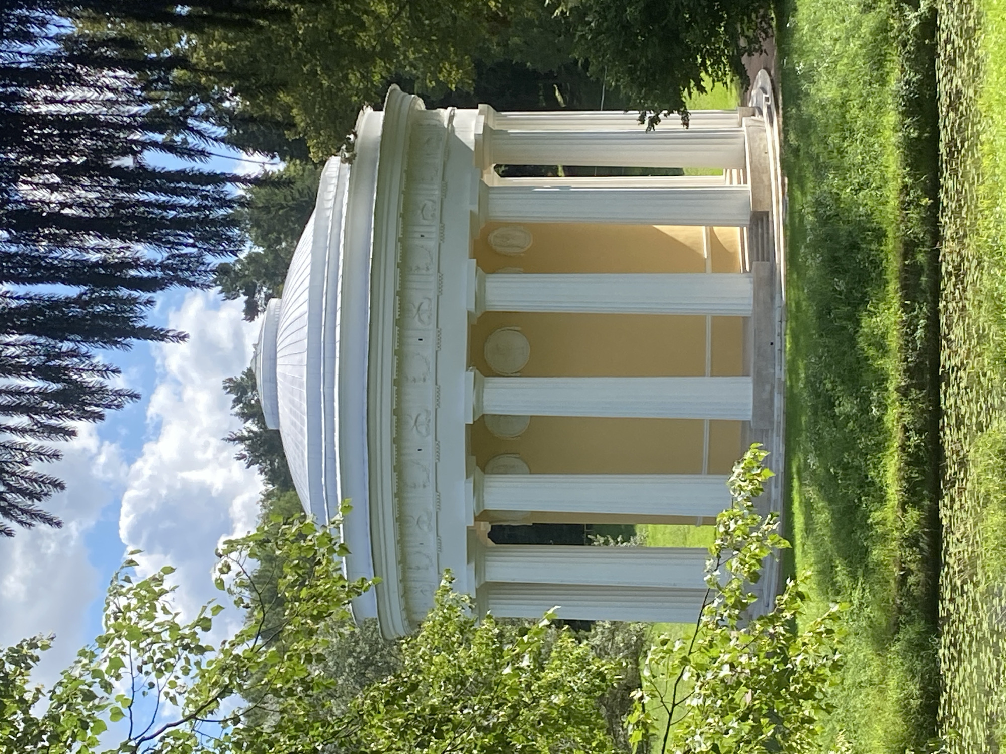 The image shows a rotunda with a white domed roof and a circular peristyle of Ionic columns. The rotunda is painted yellow and sits on a raised platform with steps leading up to it. It is surrounded by lush green grass and trees. In the foreground, there is a body of water covered with lily pads. The sky is blue with scattered white clouds.
This is the Temple of Apollo, located in the Catherine Park of Pavlovsk, Russia. The Temple of Apollo is a neoclassical pavilion built in the late 18th century. It is one of the main attractions of the park and a popular spot for tourists and locals alike.