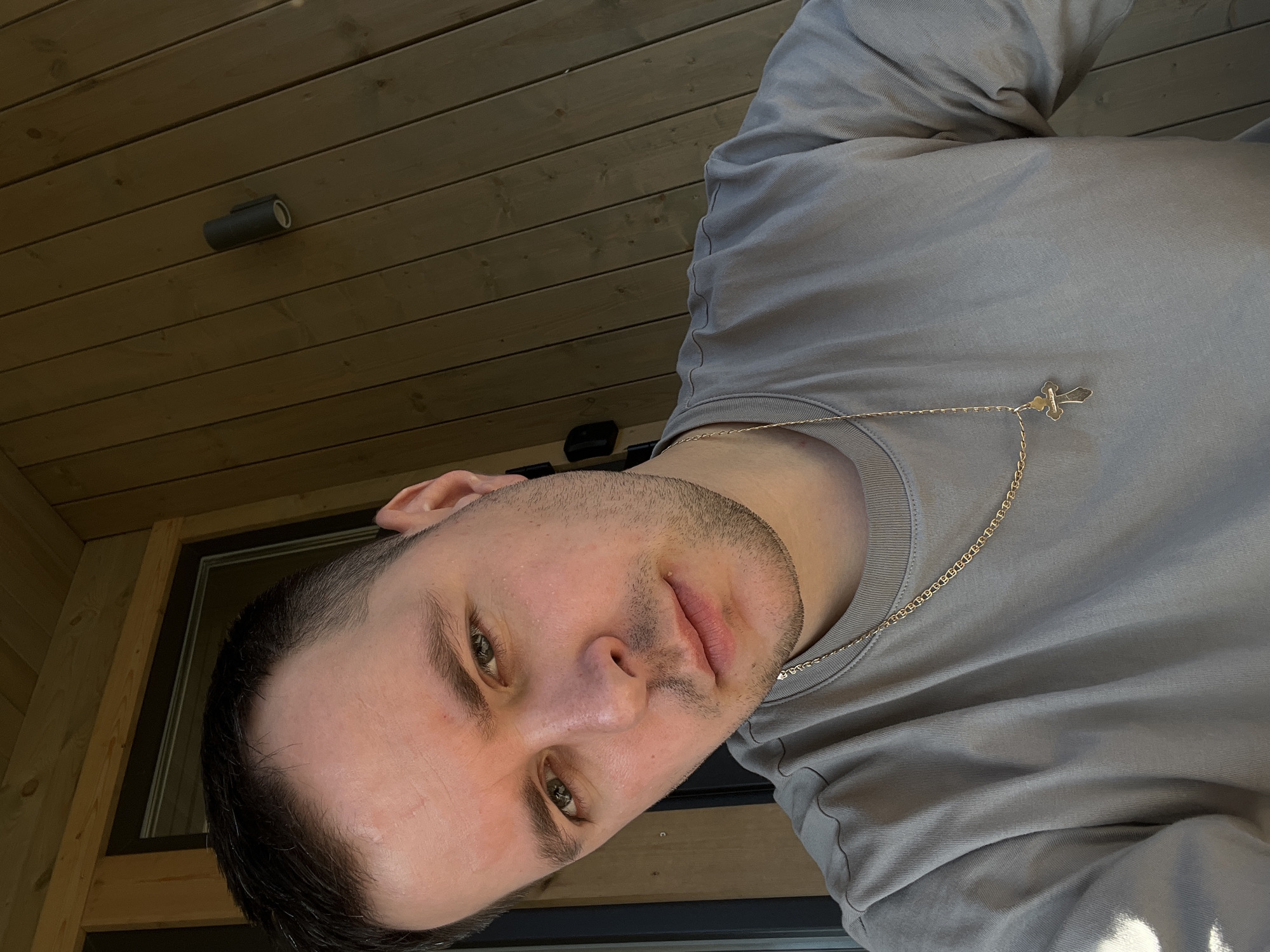 A man with dark hair and stubble is wearing a grey long-sleeved shirt and a gold chain with a small cross pendant. He is positioned in front of a window, with a wooden ceiling and an exterior light fixture visible behind him. This image does not contain enough data to determine the location to be Литвиново, Russia.