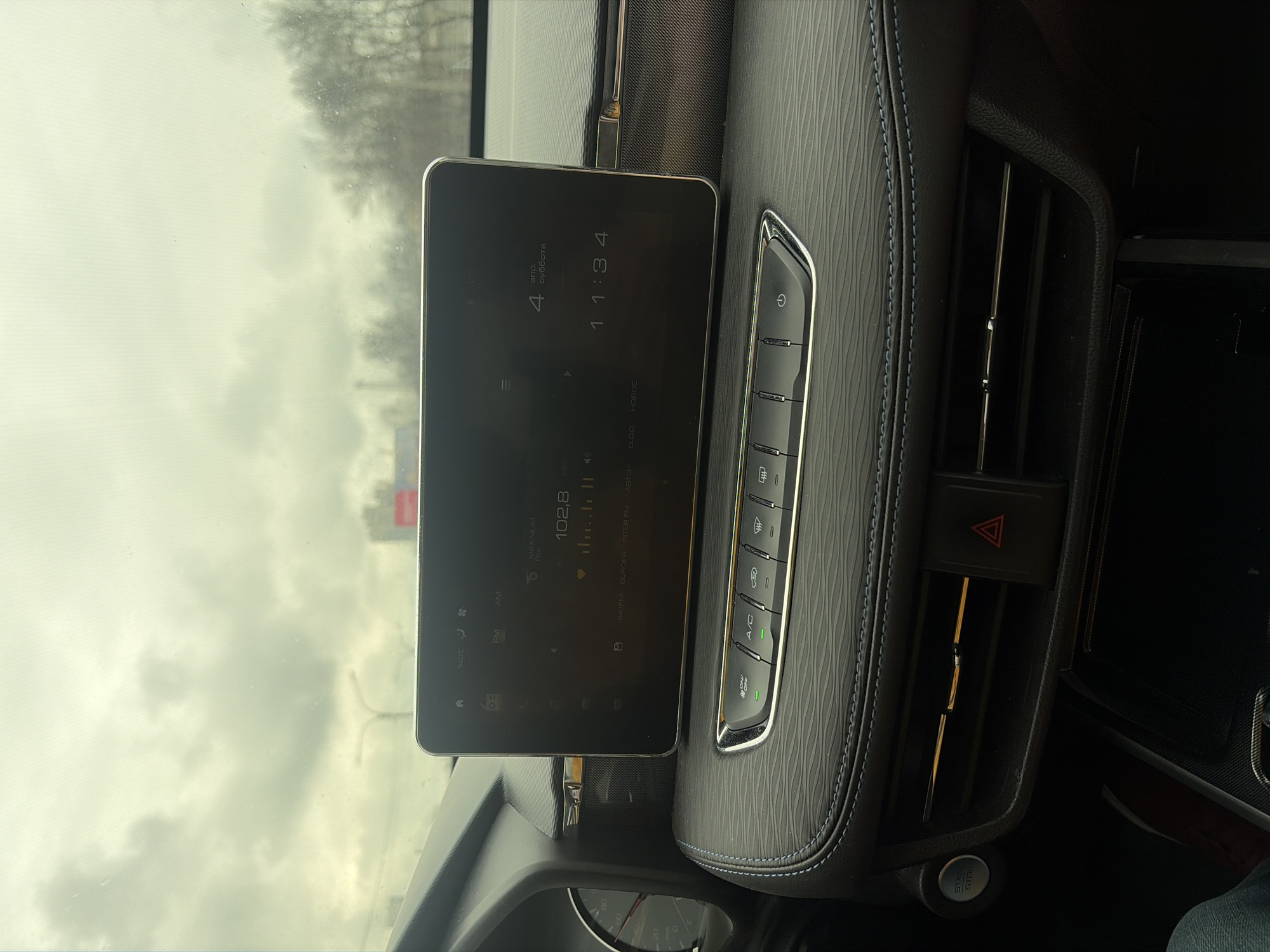 The image captures the interior of a vehicle. A central infotainment screen displays "11:34" and "102.8" on the screen. A row of buttons controls air conditioning and other vehicle functions. An air vent with a hazard light button is located nearby. The environment outside the car appears overcast. The location is unknown.