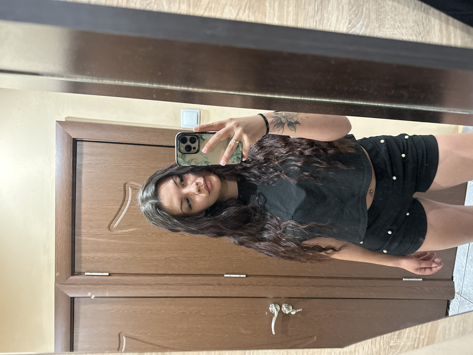 A young woman with long, dark, wavy hair is taking a selfie in a room. She is wearing a black crop top and black shorts adorned with pearl-like embellishments. Her left arm, visible in the reflection, has a tattoo of a lotus flower. She is holding a smartphone with a floral case in her right hand. The background shows two wooden doors with ornate carvings and metallic door handles. To the right of the doors, there is a glimpse of a mirror reflecting the woman and a light-colored wall. The floor appears to be tiled. The environment suggests an indoor setting, possibly a home or a hotel room in Sliven, Bulgaria.