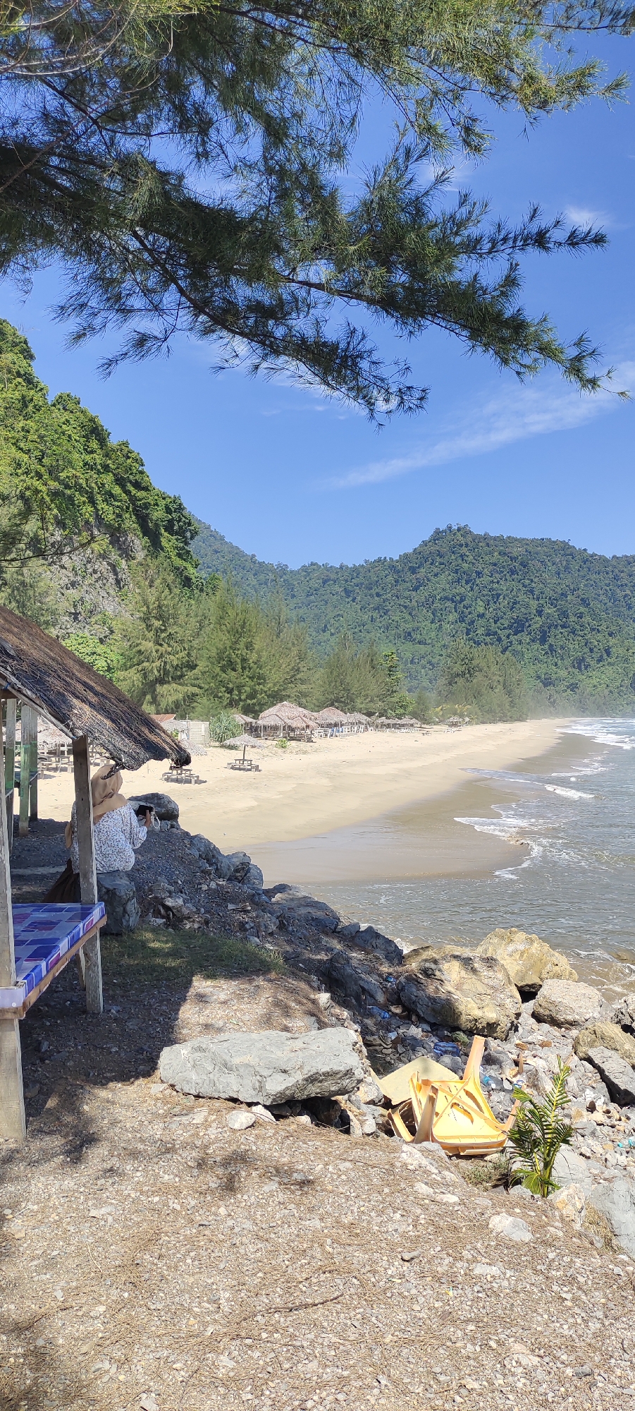 A beach scene in Meudheun, Indonesia, is captured from under a thatched-roof overhang. In the foreground, a person wearing a headscarf and a patterned shirt is seated on rocks, holding a camera. To the left, a table with a blue and white patterned surface is visible. The beach itself features a strip of sand lined with small thatched huts and palm trees. Beyond the beach, lush green hills rise under a clear blue sky with some scattered clouds. The ocean is visible on the right, with gentle waves lapping at the shore. The ground in the immediate foreground is covered in gravel and fallen pine needles. A yellow plastic chair and some scattered debris are present on the rocks to the right.