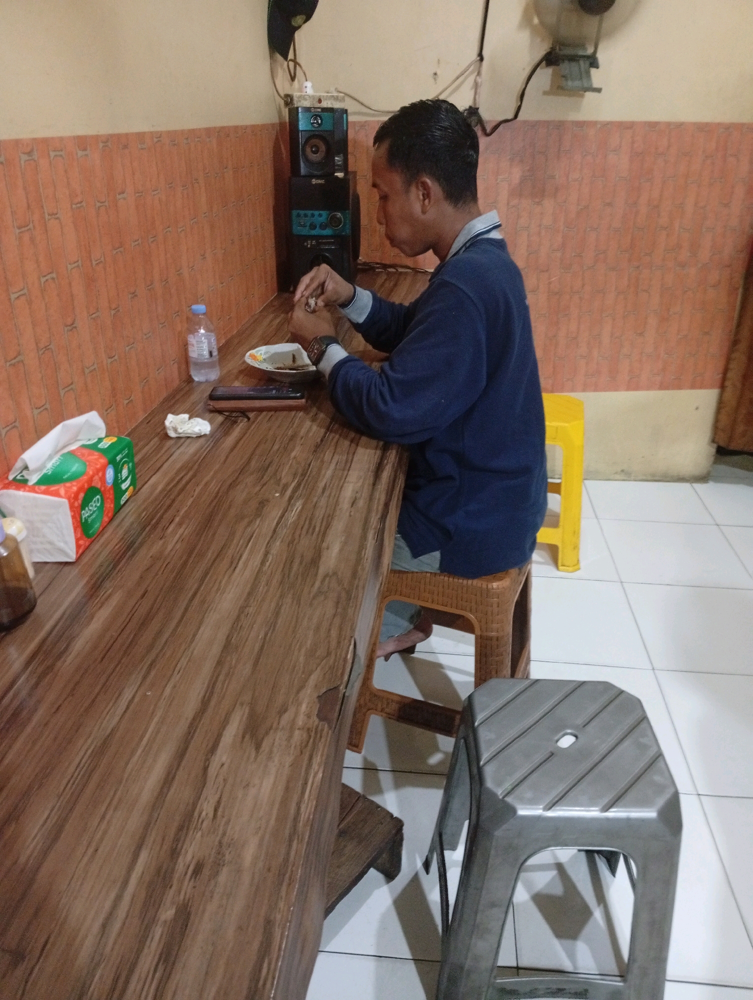 A medium shot captures a man seated at a wooden table, appearing to eat. He is wearing a dark blue long-sleeved shirt and light-colored trousers. The table is laden with items including a tissue box, a bottle of water, a plate with food, and a mobile phone. Behind the man, stacked speakers are visible against a textured orange wall. The floor is tiled in white, and a bright yellow stool and a grey plastic stool are present near the table. The scene suggests a casual dining or resting spot, potentially in a local eatery or a home in East Jakarta, Indonesia.