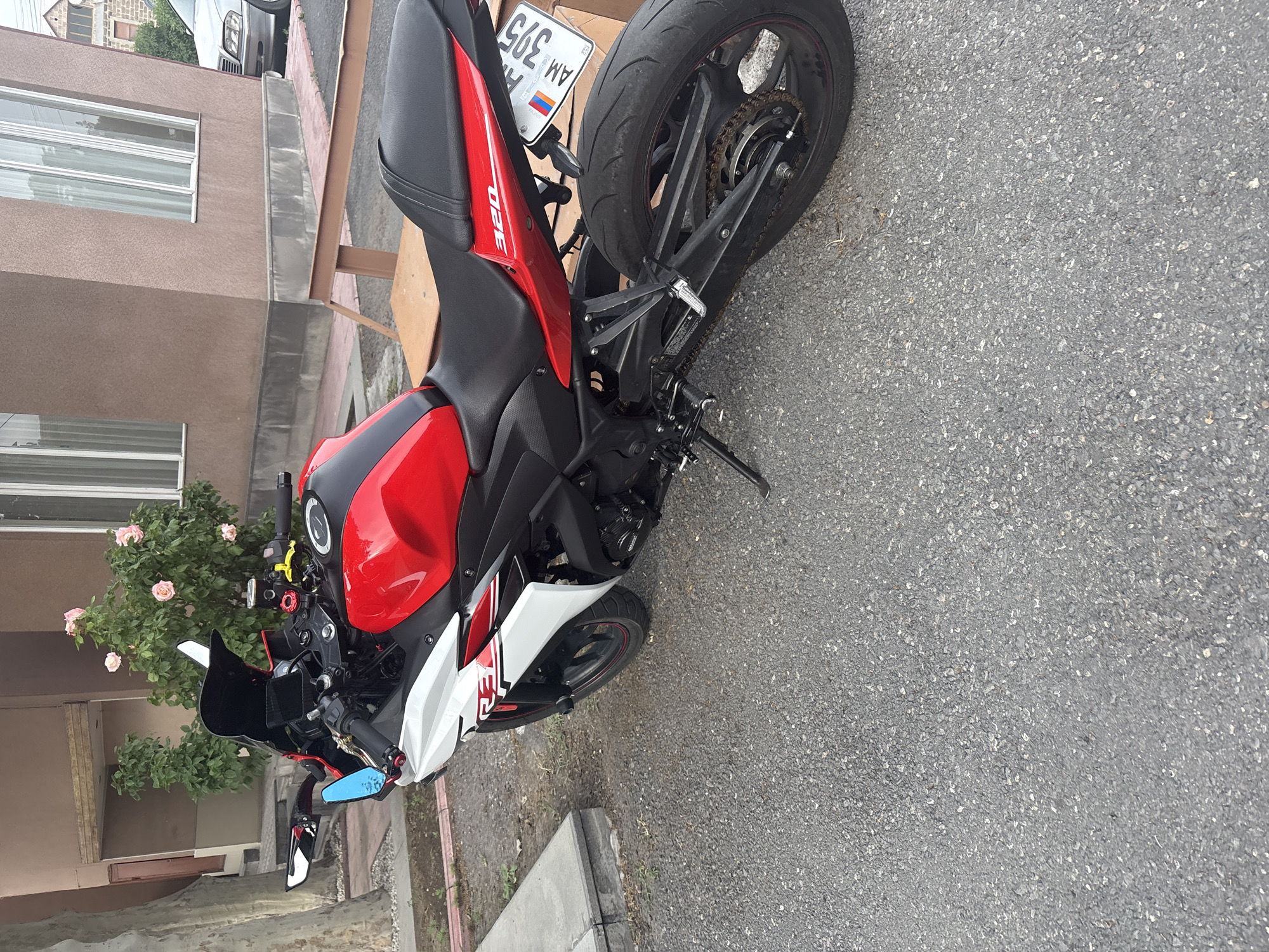 The image shows a red and white Yamaha 320 motorcycle parked on an asphalt street. The motorcycle's license plate reads "A.M. 395," indicating it is likely in Armenia. The red, blue, and orange flag design on the license plate is a match for the Armenian flag. The presence of what seems to be a flower bush and a building in the background suggests an urban or residential area. Based on the license plate and other location clues, the location is likely Yerevan, Armenia.