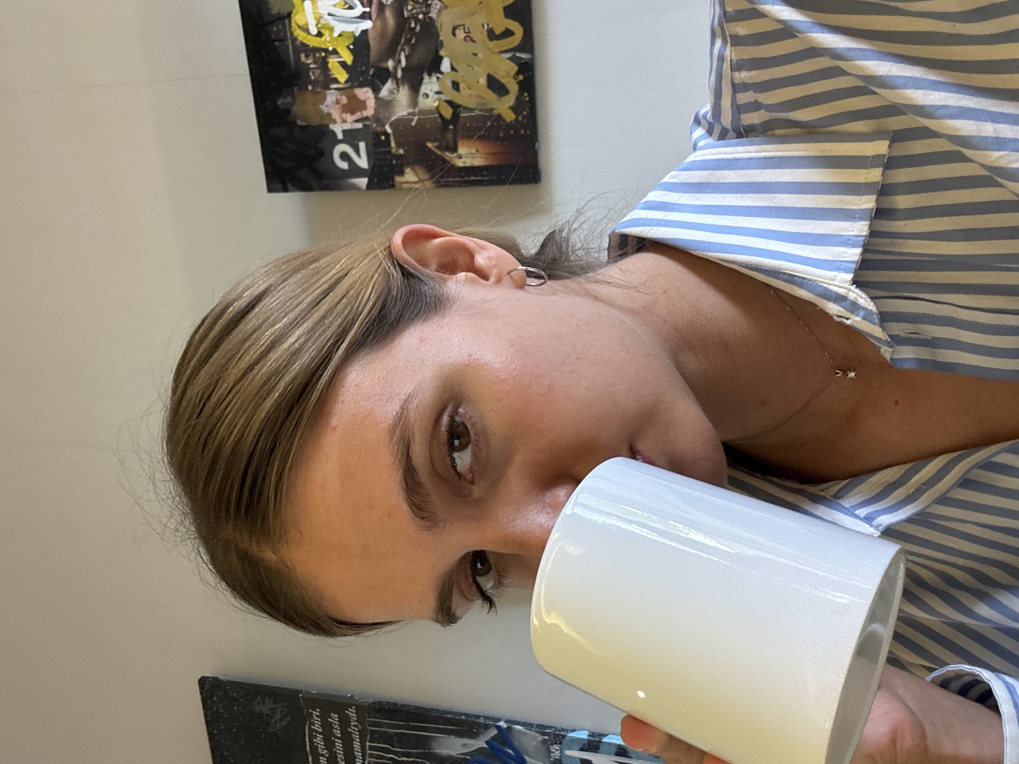 The image is a medium close-up, angled portrait of a person holding a white mug. The individual, with light brown hair, is seen from the chest up, seemingly looking towards the viewer while bringing the mug to their lips as if to drink. They are wearing a blue and white striped button-up shirt.
Notable details include a small silver hoop earring in the helix of their right ear and a delicate silver chain necklace with a tiny, possibly diamond or gemstone, pendant. The person's expression is neutral, and their eyes are a light brown color.
The background consists of a plain, light-colored wall. Above the person's head, an abstract artwork or poster is partially visible. This piece features yellow squiggles on a dark background, with the number "21" clearly discernable, albeit rotated due to the image's orientation. Below the person's chin, a dark object resembling a book or piece of paper is visible, containing text in what appears to be Turkish: "um gibi biri, esini asla namalıydı."
The scene is set indoors, suggesting a casual environment like a home or office. No specific time of day or further location cues are evident.