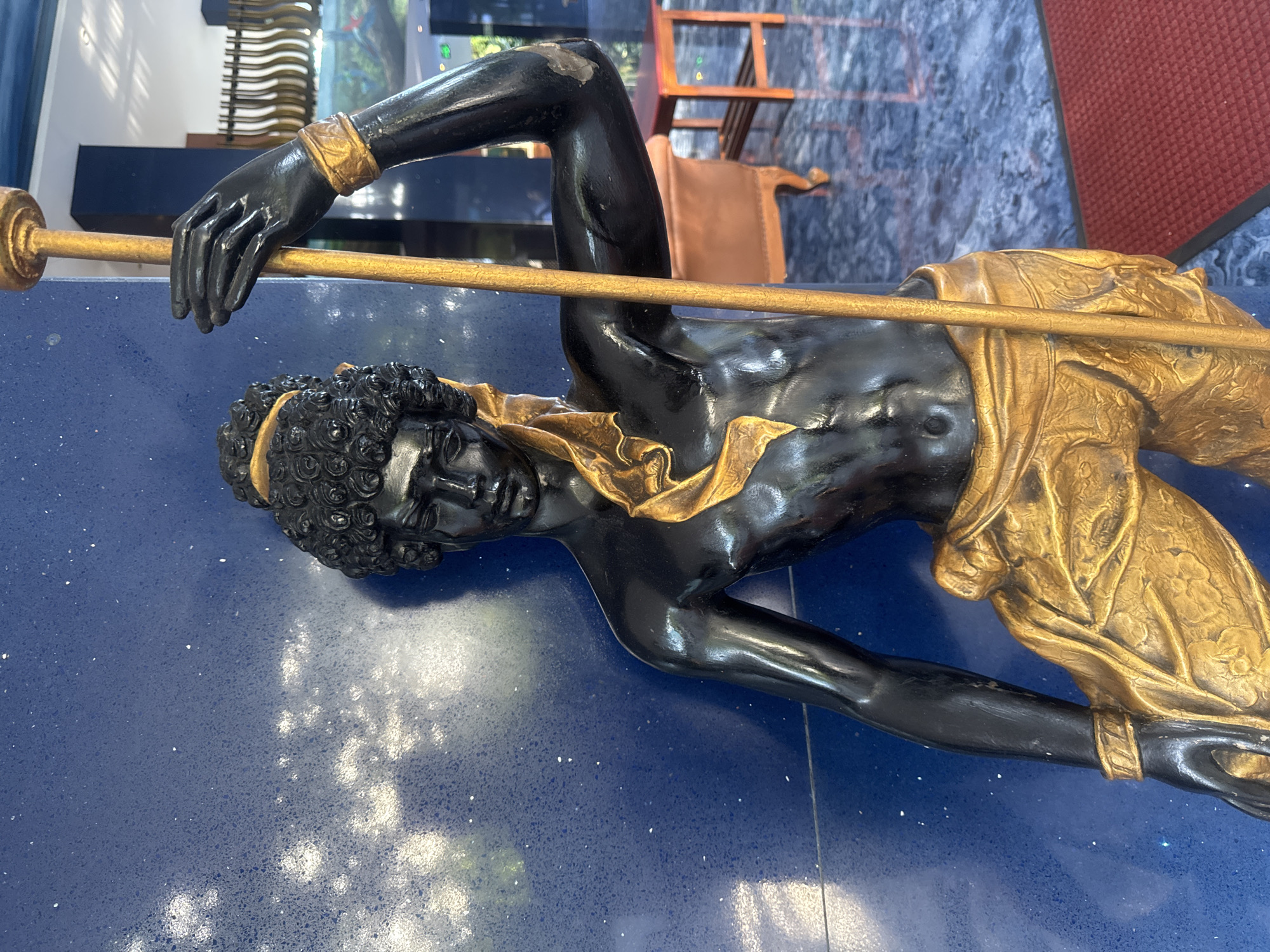 A statue of a black figure adorned with gold elements lies diagonally across a blue, speckled surface. The figure has curly hair, a gold headband, and a gold draped garment around its lower body. The figure appears to be holding a long gold pole. The background is slightly blurred, but a chair and other furniture pieces can be seen. This statue is located in Kuta, Indonesia.