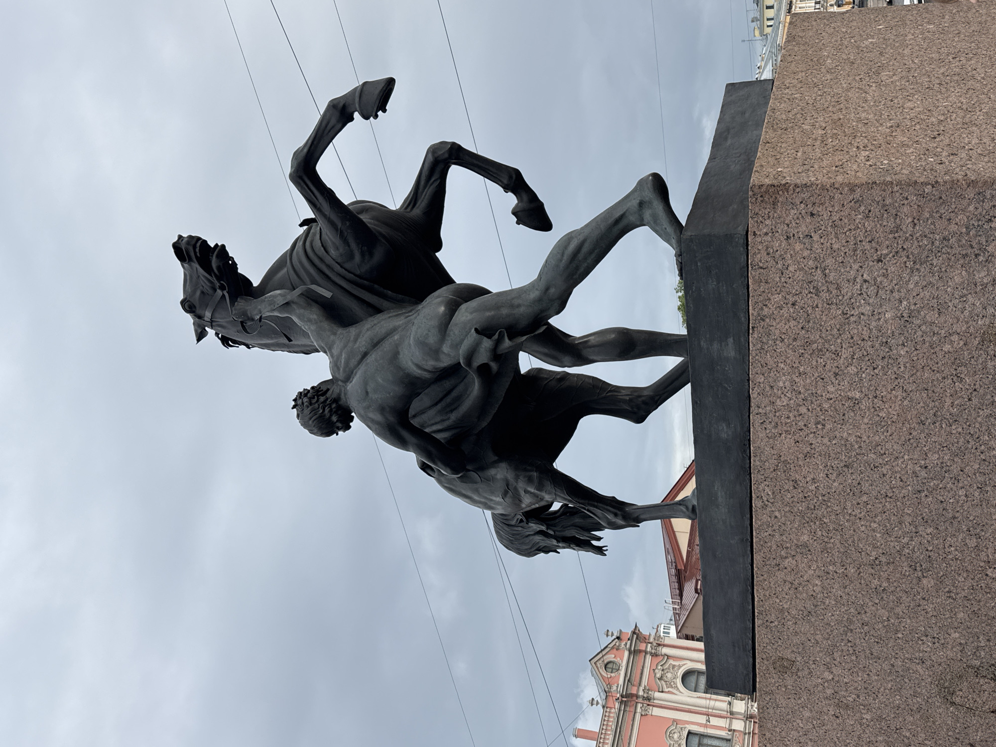 This is a low-angle, vertical shot of a bronze sculpture depicting a man wrestling with a horse. The sculpture is positioned on a stone pedestal, with a dark, textured surface forming the base. The man is shown in a dynamic pose, his body entwined with the horse's, appearing to struggle for dominance. The horse is rearing up, its hind legs in the air, with its head turned downwards. The musculature of both figures is clearly defined, emphasizing the tension and power of the struggle.
In the background, a cloudy, overcast sky dominates the upper portion of the image. Below the sculpture, a pink building with ornate architectural details and arched windows is visible, suggesting an urban setting. The overall mood is one of intense effort and dramatic conflict. The lighting suggests it is daytime, but the overcast sky lends a subdued atmosphere. The composition, with the sculpture filling most of the frame and extending upwards, emphasizes its grandeur and imposing presence. The sculpture is likely an equestrian monument or a representation of a mythological or historical scene.