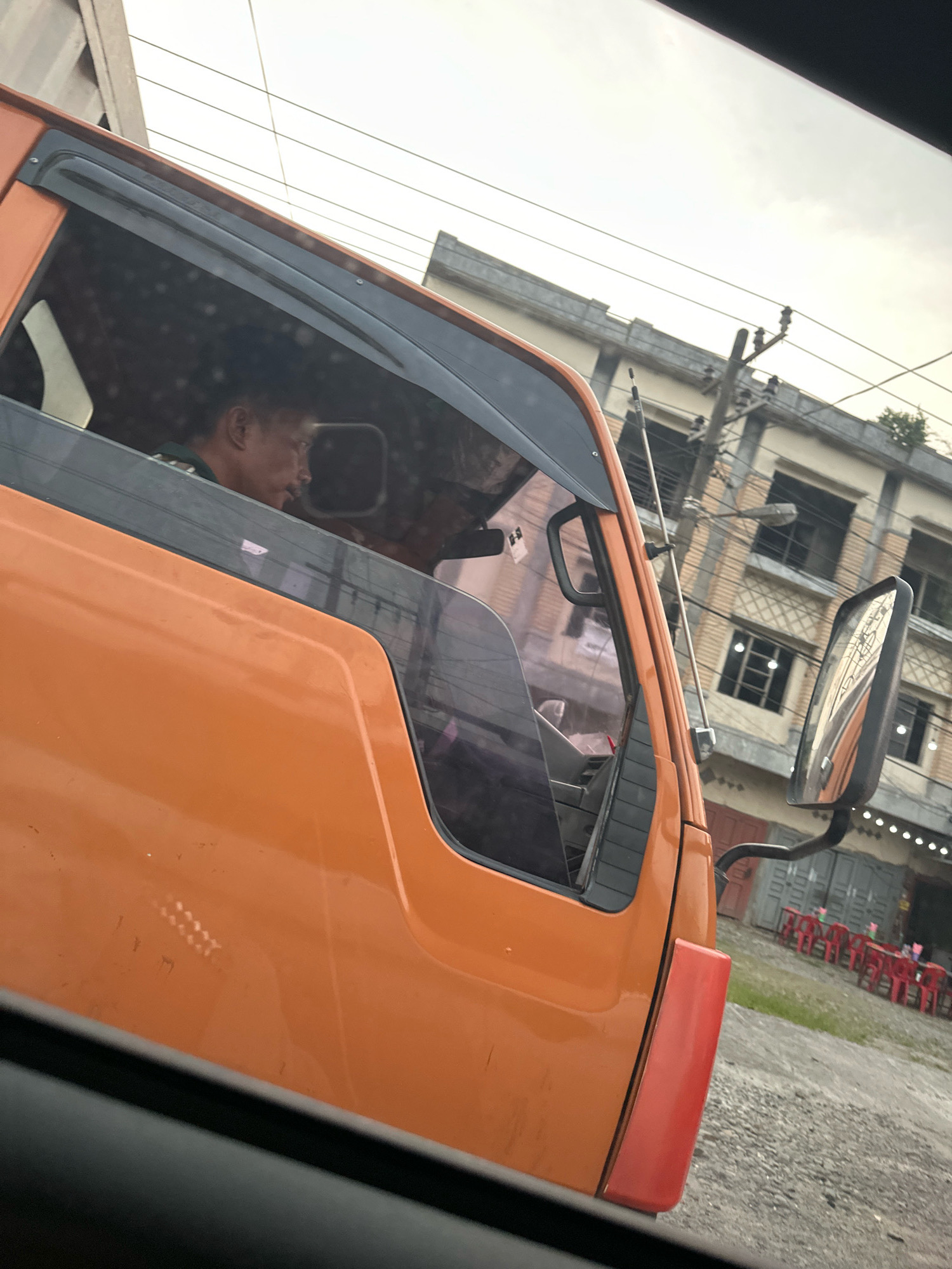 An exterior, eye-level, slightly tilted, medium shot captures the driver's side of an orange truck. The driver, a man with dark hair and a mustache, is visible through the tinted window, looking to his right. The truck's large side mirror reflects a building with multiple floors and windows, appearing to be in an urban setting with power lines overhead. In the background, a row of red chairs and tables are visible outside a building, suggesting an outdoor dining area. The ground is covered with gravel. The sky is overcast. The overall scene appears to be in the City of Binjai, Indonesia.
