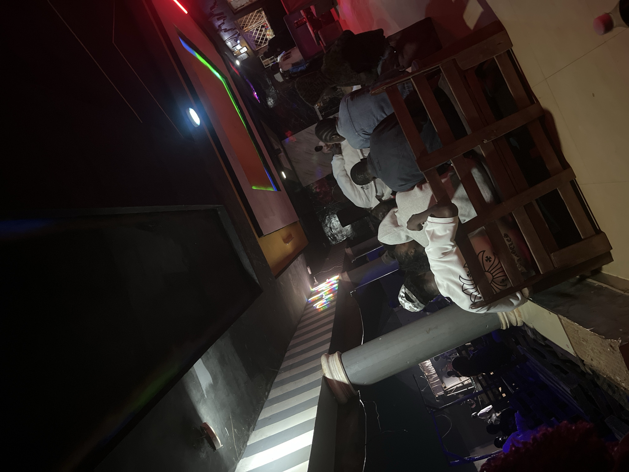 The image depicts a dimly lit indoor scene, likely a bar or lounge, with a group of people seated. In the foreground, several individuals are seen from behind, facing away from the camera. They are seated on wooden chairs, suggesting a communal seating arrangement. The lighting is atmospheric, with colored lights illuminating the ceiling and walls, creating a vibrant and possibly energetic ambiance. In the background, other patrons are visible, some standing and others seated, further indicating a social gathering. The decor appears modern with textured walls and ceiling elements, possibly featuring striped patterns. The overall impression is of a lively establishment, likely a nightlife venue. Based on the context provided, this scene is located in Durimi, Nigeria.