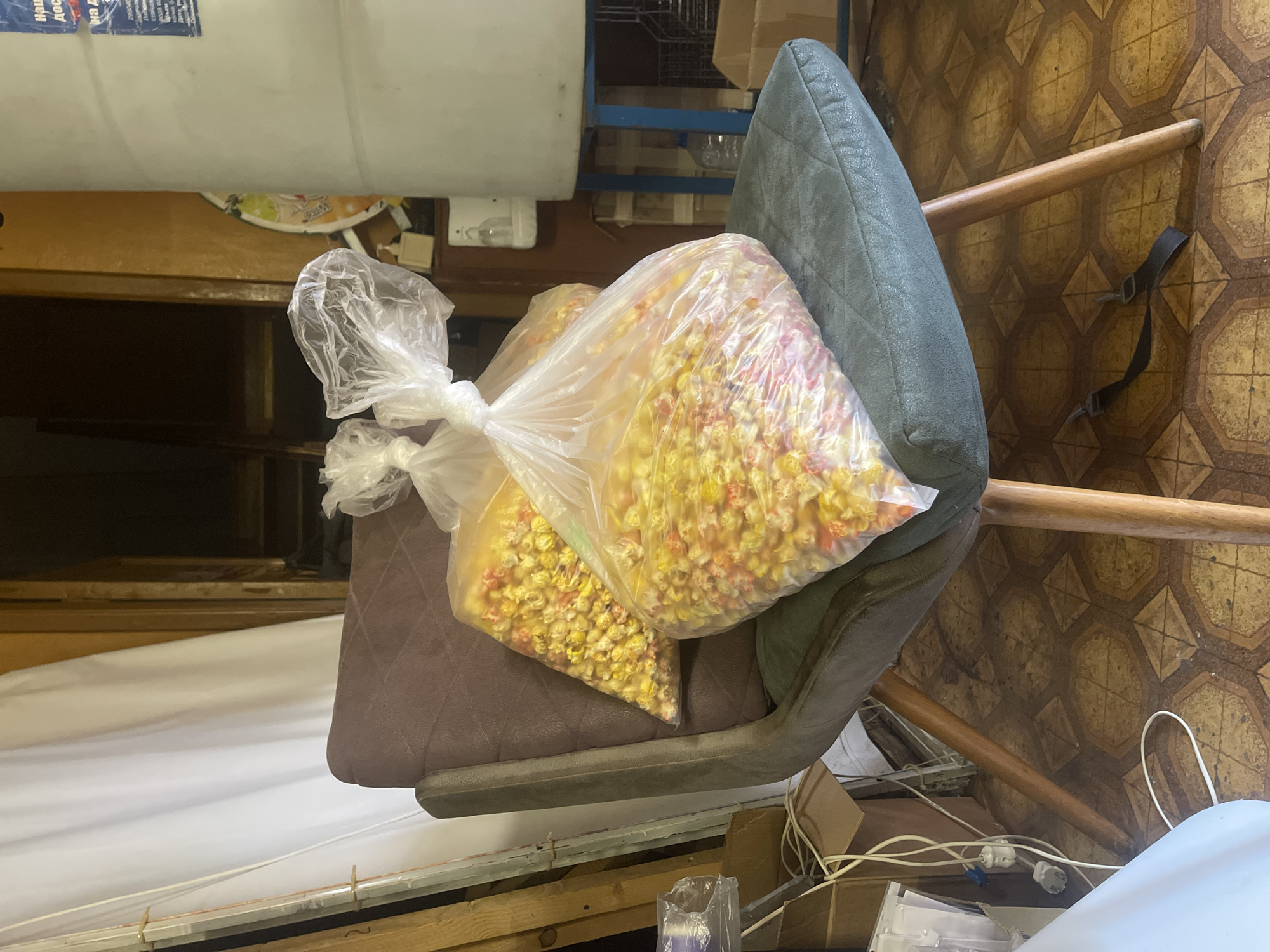 An indoor scene within the city of Unknown, Unknown, displays two large, transparent plastic bags of multi-colored (yellow, red, and white) popcorn resting on a dark gray upholstered chair with wooden legs. The chair features a quilted pattern on its back and seat. To the right, a tall, white cylindrical tank, supported by a blue metal stand, is visible, with a partially obscured label featuring Cyrillic text on its upper section. Below the tank, a small white wall-mounted dispenser is affixed to a wooden cabinet. The floor is covered with an old, patterned linoleum or tile featuring octagonal and square shapes in brown and yellow tones. On the left side of the image, various items are present, including a tangle of white electrical wires and extension cords, a blue plug, cardboard boxes, and several wooden planks leaning against a wall. A white curtain hangs along the left wall. A black fabric strap with two clips lies on the floor beneath the chair. The background on the right shows a wooden doorway leading to a darker, indistinct area.