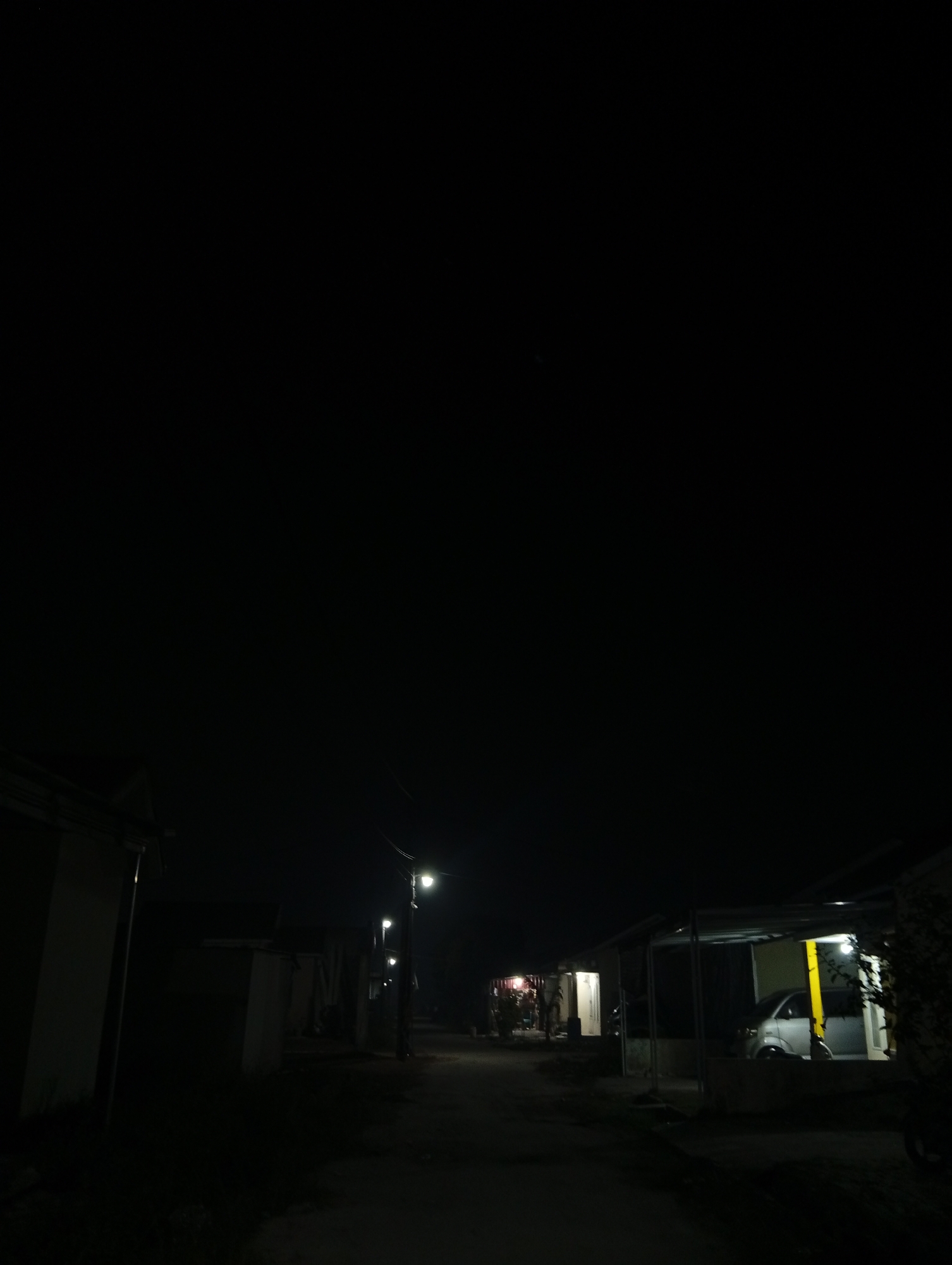 The image captures a nighttime street scene in Kualu, Indonesia, characterized by darkness punctuated by artificial lighting. Several low-lying structures, likely homes or small businesses, line a narrow dirt road. One building on the left has a light-colored facade. Further down the road, streetlights illuminate the path and cast shadows on the surrounding buildings. On the right side of the image, a silver vehicle is parked under a covered carport, with a bright yellow wall visible behind it. Another structure further down the street has a red glow emanating from it. The overall atmosphere is quiet and still, with no visible people or animals. The environment appears to be a residential or semi-rural area.