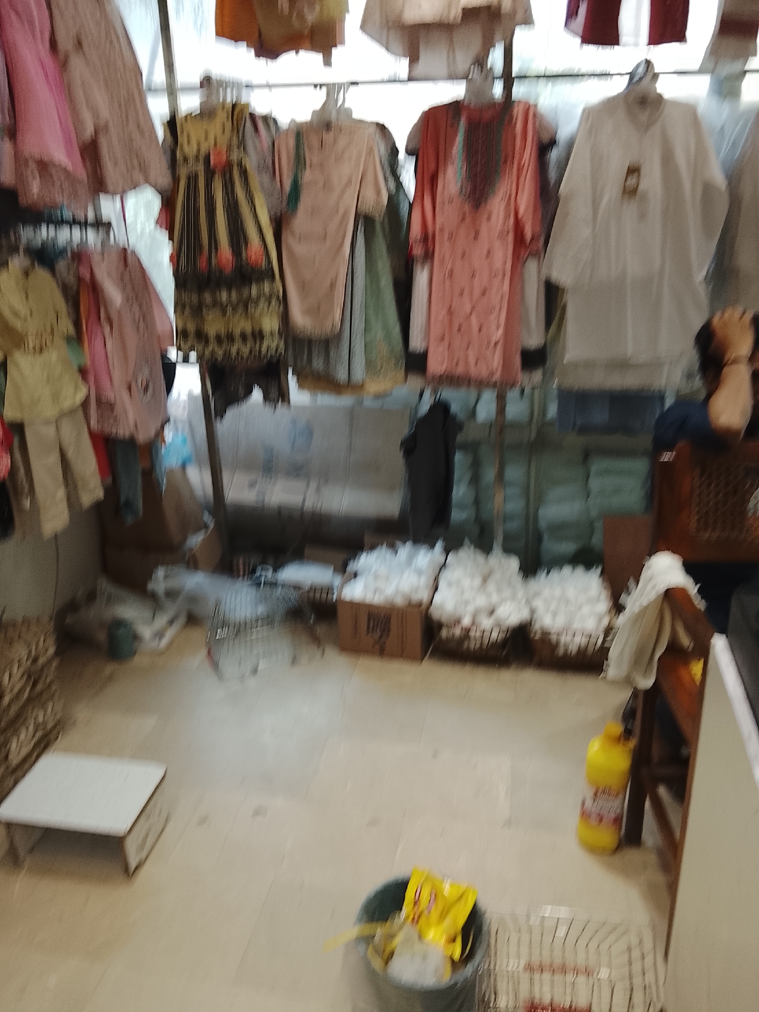 The image depicts a retail space in Gulgasht Colony, Pakistan, filled with clothing items displayed on hangers and racks. The clothing includes various garments such as dresses, shirts, and children's outfits, in different colors and patterns. Below the clothing racks, there are baskets and boxes filled with what appear to be cotton products or textiles. Other objects in the scene include a small white stool, a bottle of yellow liquid detergent, a waste bin containing trash, and shelving. The environment appears to be a commercial area, possibly a store or market, characterized by a cluttered arrangement of merchandise.