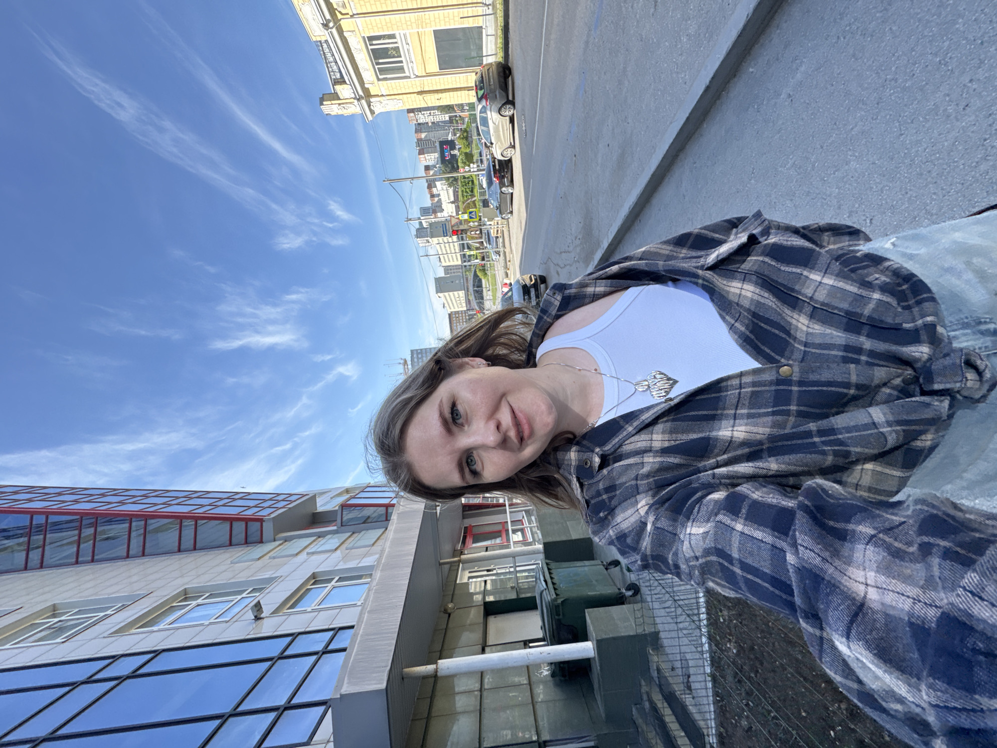 An outdoor selfie captures a young woman in an urban setting, rotated 90 degrees counter-clockwise. The woman is positioned in the foreground, smiling gently towards the camera. She has light brown hair, blue-green eyes, and is wearing a white tank top under an unbuttoned blue and beige plaid shirt. A silver heart-shaped pendant hangs from her neck.
The background reveals a bustling city environment under a bright, clear blue sky with wispy white clouds, suggesting a sunny daytime. To her left (if the image were upright), a modern building with large reflective glass windows and red accents is visible. To her right, a street or sidewalk features parked cars and another building with a more traditional, possibly yellow-hued facade. Further in the background, a cityscape with various buildings and structures extends into the distance. The scene depicts a typical city street, with no discernible text visible. The overall mood is relaxed and pleasant. The location context is Perm, Russia.