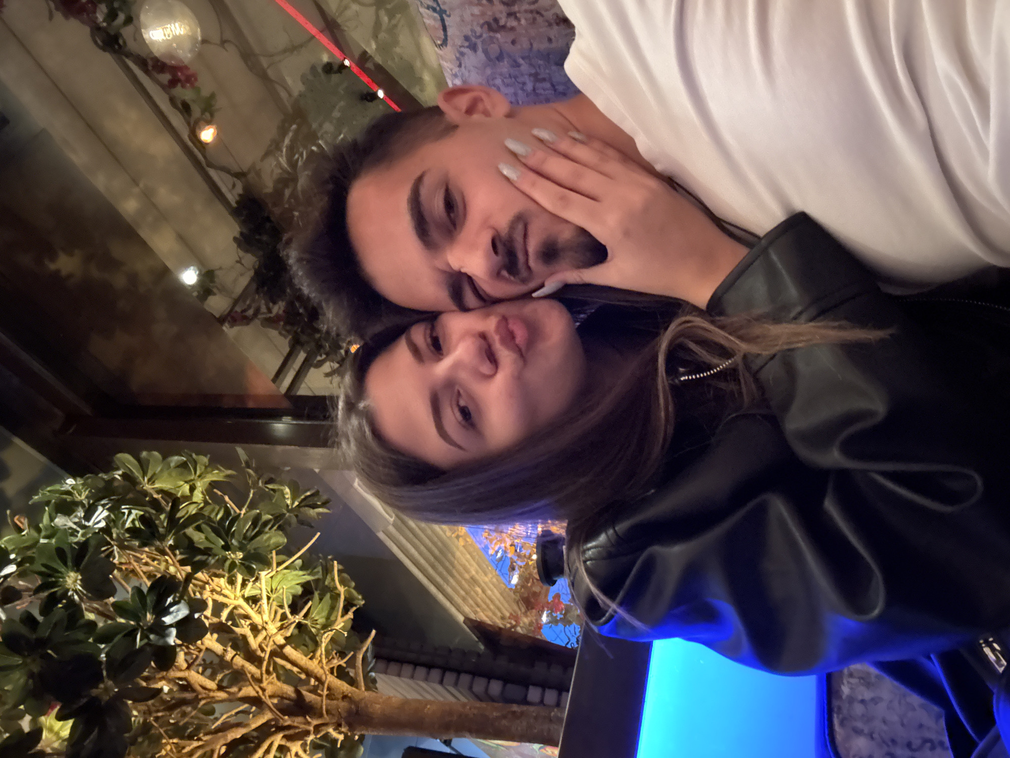 The image captures a close-up selfie of two individuals, a female and a male, in an indoor establishment, likely a dimly lit restaurant or bar, located in Sofia, Bulgaria. The female, positioned on the left, has long dark hair, wears a black leather jacket, and poses with pursed lips in a kissing gesture directed towards the male. Her left hand gently cups the jawline of the male subject. The male, on the right, has short dark hair, a mustache, and a goatee, and wears a plain white t-shirt. He squints playfully, making a humorous, slightly scrunched expression.
The background on the left features a decorative artificial tree with a textured brown trunk and green foliage, warmly illuminated from below by a yellowish light. Below the tree, a bright blue ambient light source is visible. The upper background, particularly on the right, reveals a reflective ceiling (possibly mirrored) adorned with draped artificial vines, small warm incandescent bulbs, and hints of red decorative elements, creating an ornate and cozy ambiance. A patterned, upholstered chair or booth seat in muted tones is visible behind the male on the lower right. The scene conveys a moment of casual intimacy and playful interaction.
