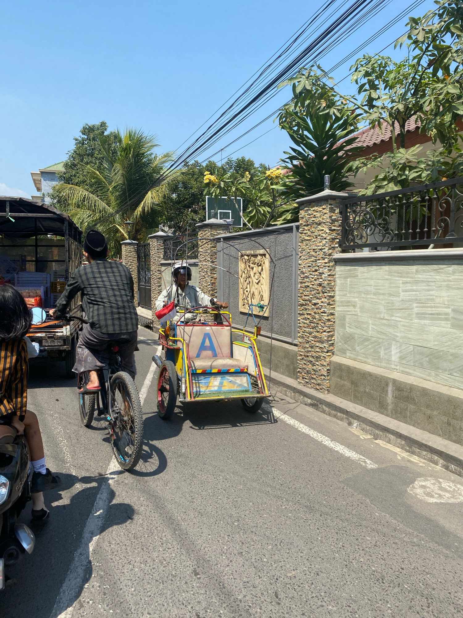 In Kediri City, Indonesia, a bright, sunny day showcases a street scene with various modes of transportation and local architecture. In the foreground, a person on a bicycle, wearing a plaid shirt and a peci, navigates the road. To their right, a colorful pedicab with a large blue "A" on its side is being driven by a man wearing a helmet and holding a small Indonesian flag. Behind the cyclist, a large truck with a canopy is visible, loaded with goods. The roadside features a stone-clad wall with an ornate metal fence and lush greenery. The road itself is asphalt with white lane markings, and a visible drainage ditch runs along the curb. The background reveals more residential buildings and tall trees, with a clear blue sky overhead.