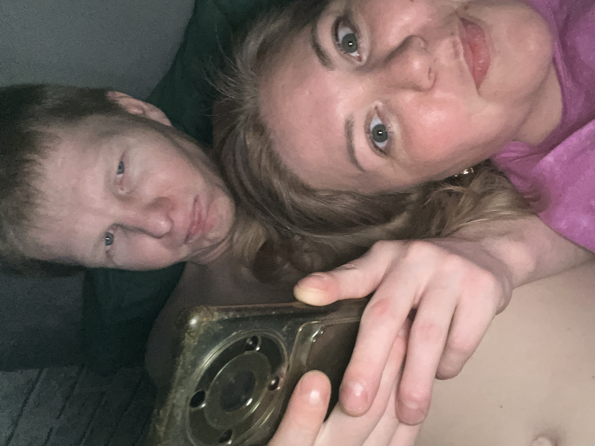 The image shows a selfie taken by a woman with fair skin and light brown hair, wearing a pink top. A man with fair skin and light red hair is also in the frame. They are holding a mobile phone with a worn, dark-colored case. The background appears to be a dark green or gray surface. The location, Vesyoloye, Russia, is not discernible from the image.