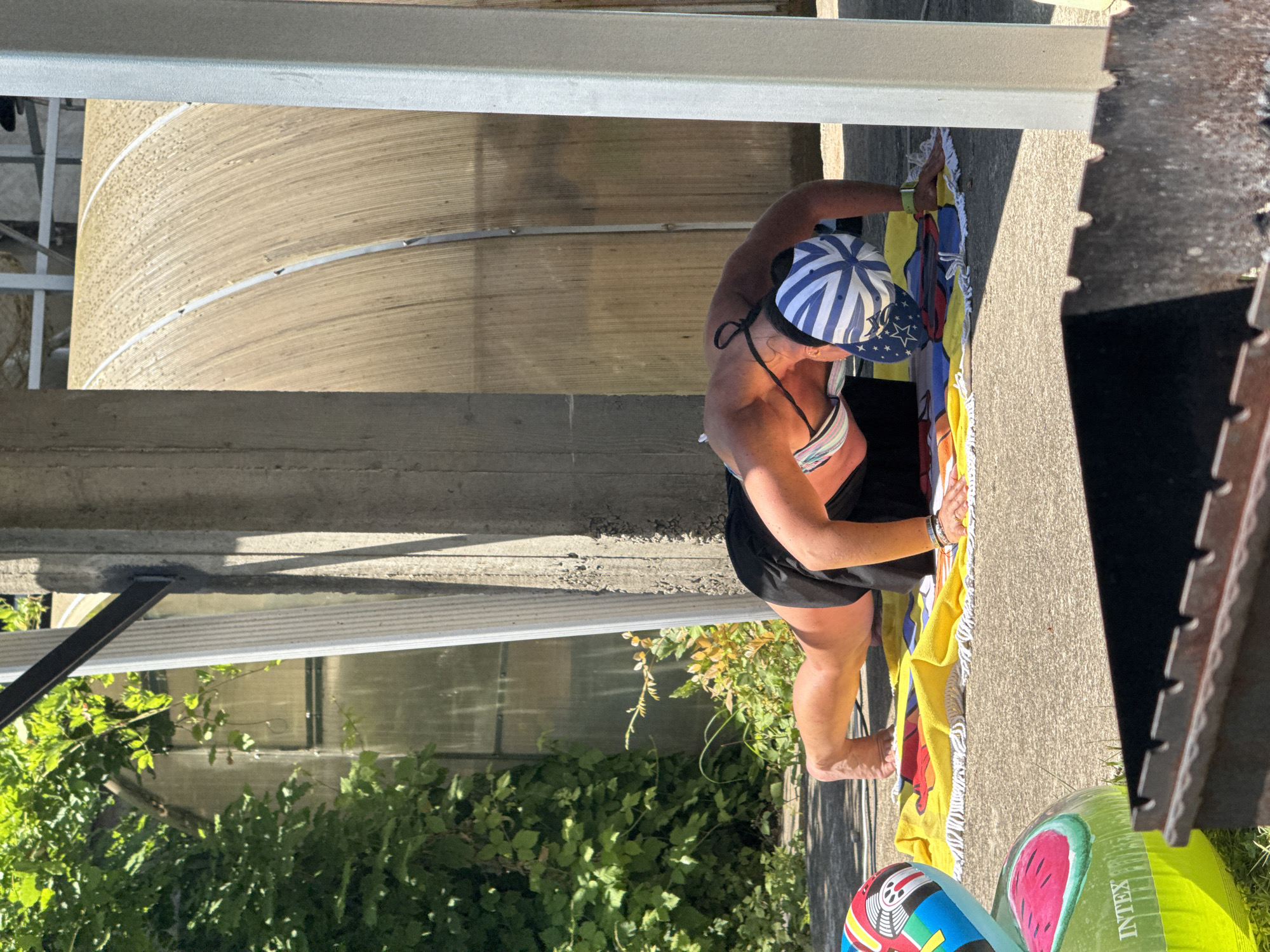 A woman is doing push-ups on a colorful towel laid out on a paved surface, possibly a patio or driveway, in Sochi, Russia. She is wearing a black athletic skirt, a striped bikini top, and a blue and white striped baseball cap with stars. Green foliage is visible in the background, along with a concrete structure and a large, curved, metallic structure. In the foreground, there are inflatable pool toys, including one with a watermelon design, and a dark metal barbecue grill. The scene appears to be outdoors on a sunny day.