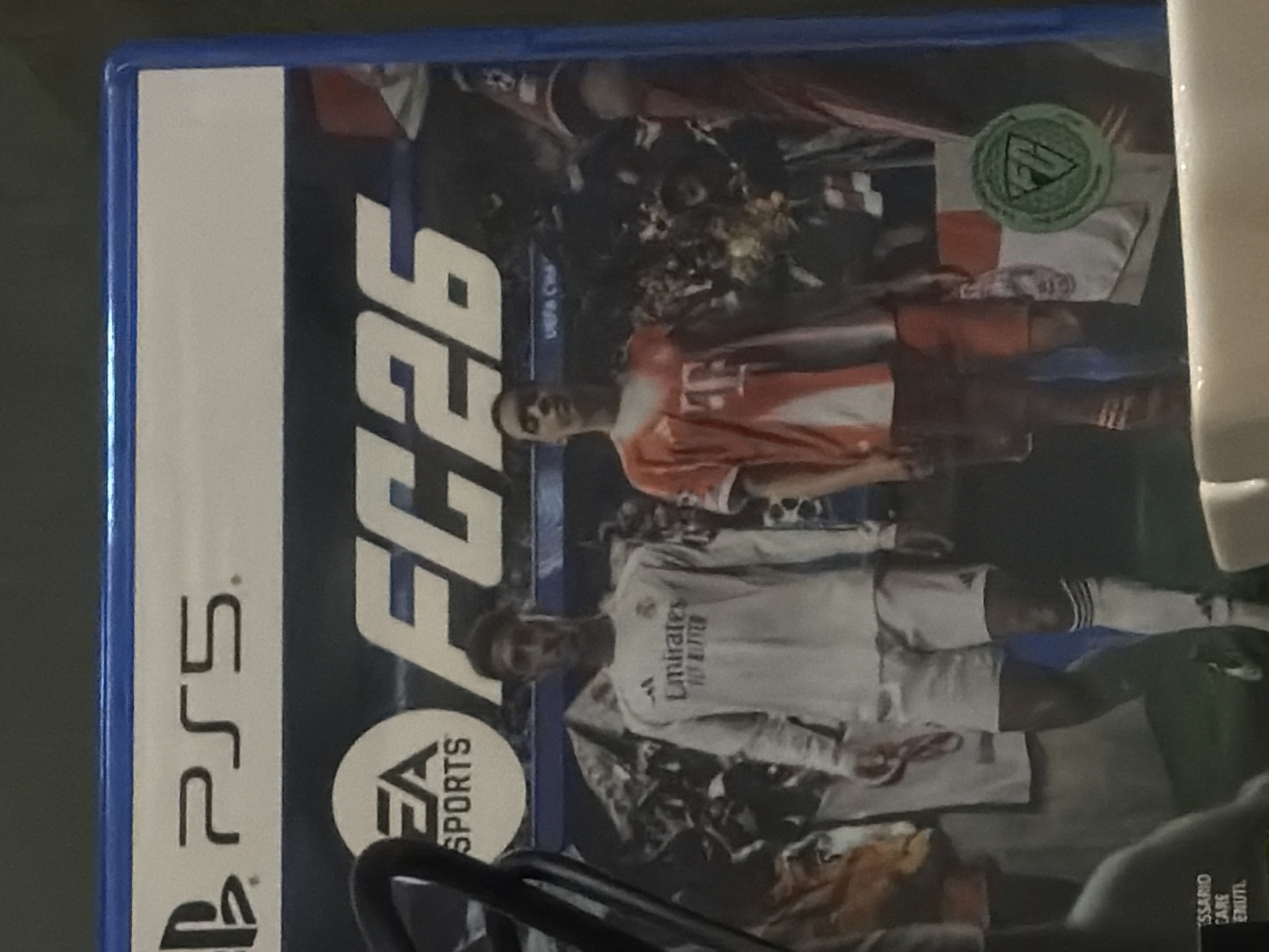 The image is a close-up of the cover art for the video game "EA SPORTS FC 26" for PlayStation 5. The cover features two male soccer players standing in front of a blurred crowd. The player on the left is wearing a white Real Madrid jersey and shorts. The player on the right is wearing a red and white jersey and red shorts. The EA SPORTS logo and "FC 26" are prominently displayed at the top of the cover. The PlayStation 5 logo is also visible in the upper left corner. There is a green circular emblem with a white "FP" inside it in the lower right corner. The background is dark and appears to be a stadium.