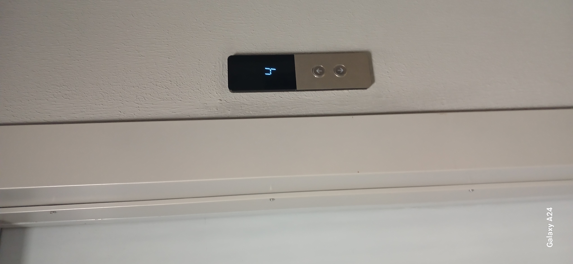 A digital display shows the number "4" on a black elevator panel attached to a textured wall. Below the digital display are two circular buttons, one with an upward-pointing arrow and the other with a downward-pointing arrow. To the left of the elevator panel is a white door frame. The bottom left corner of the image shows the text "Galaxy A24".