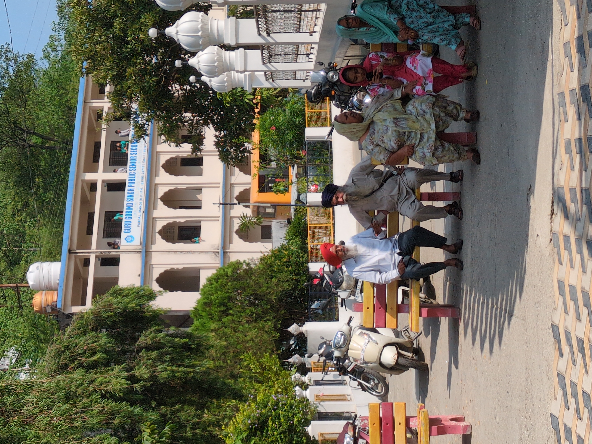 In Mandi, India, an outdoor scene depicts a group of people sitting on benches in front of a building. The building, likely a school or religious institution, has a light-colored facade with arched windows and a blue trim. Above the building, water tanks are visible. Several motorcycles and a scooter are parked nearby. The foreground features a patterned pavement and shadows cast by an unseen object. The people, including men and women, are dressed in traditional Indian attire. Some are wearing turbans. One woman is looking at her phone, while others are conversing or observing their surroundings. The environment is sunny, with trees and greenery surrounding the area. The overall impression is one of a casual gathering in a public space.