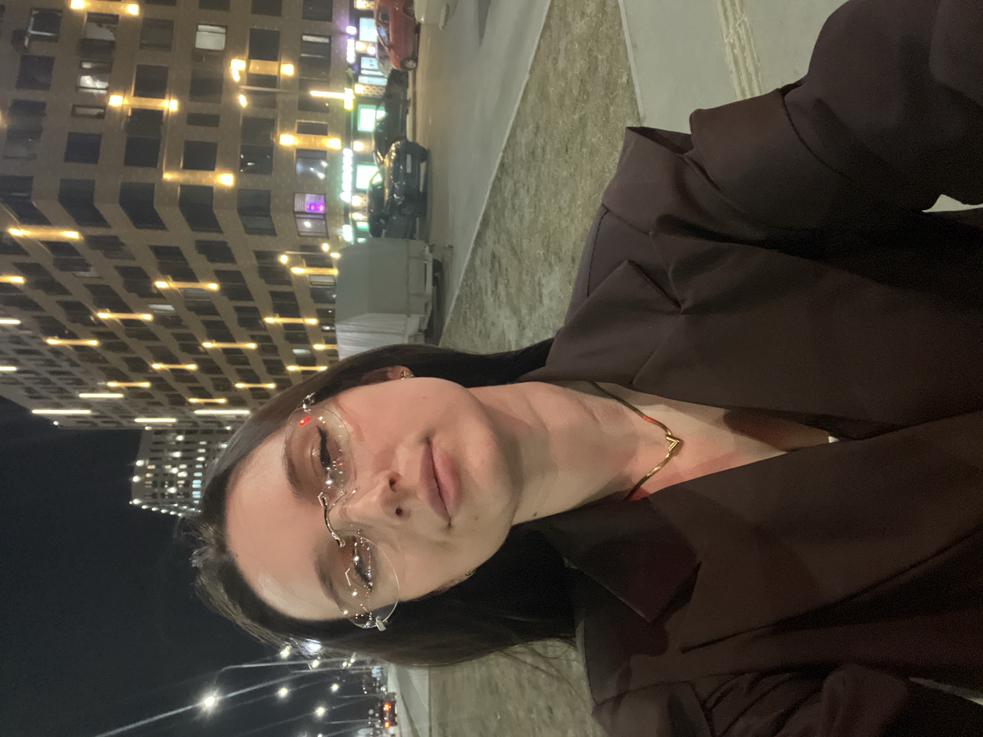 A woman is posing in a vertically oriented selfie shot at night on a city street. She is wearing glasses with clear, rimless frames and a dark brown blazer. A gold necklace is visible at her throat. In the background are city street lights, cars, and a tall building with many illuminated windows. It's difficult to provide any further details regarding the location and people in the image.
