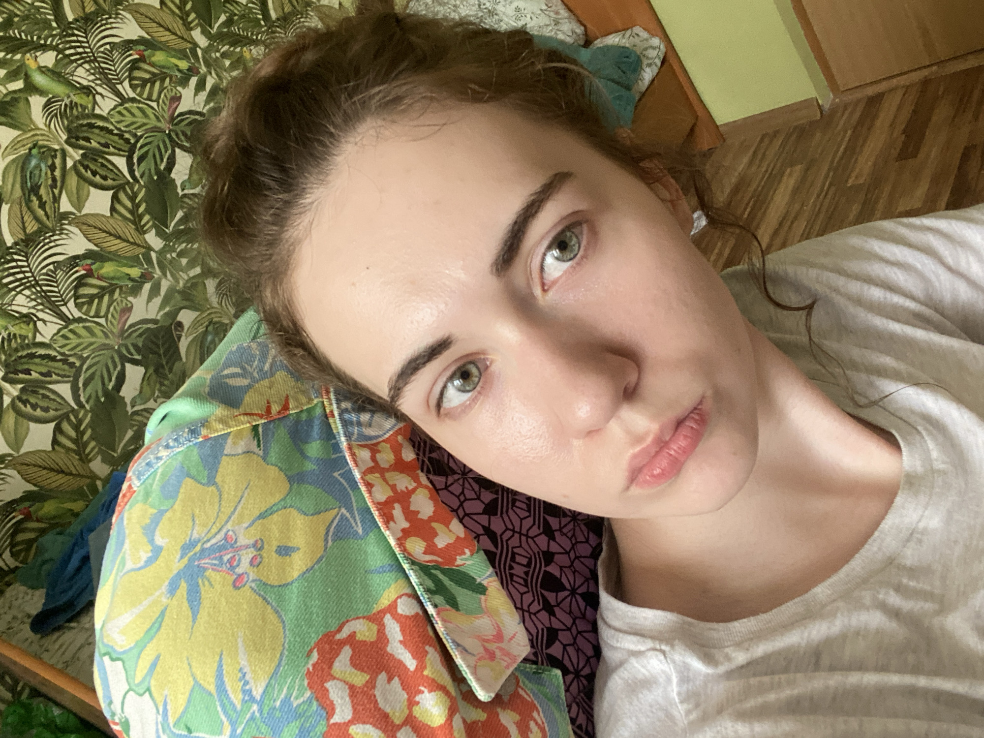 A young woman with light skin, brown hair pulled up, and green eyes is lying down. She is wearing a light gray t-shirt. Behind her, a colorful pillow with a floral and fruit print is visible. The background consists of patterned wallpaper with green leaves and birds, and wooden floorboards. The scene is indoors, likely in a bedroom. The city of Zavodoukovsk, Russia is not visually represented in the image.