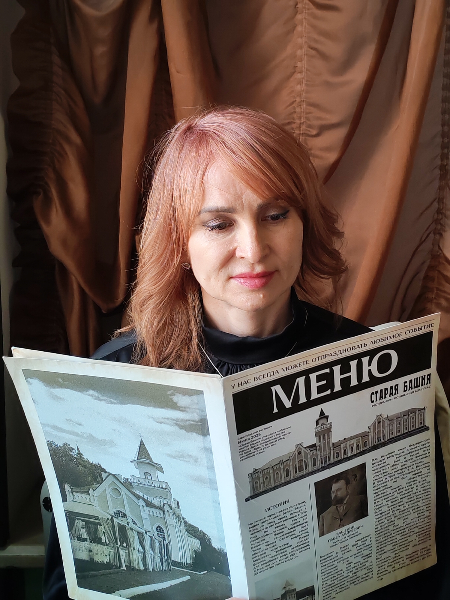 A woman with reddish-blonde hair is reading a newspaper. The newspaper appears to be a special edition focusing on the history and promotion of the "Staraya Bashnya" (Old Tower) restaurant and hotel complex in Sarapul, Russia. The open pages of the newspaper feature photographs and text about the establishment. The woman is positioned in front of a brown, textured backdrop that may be a curtain or draped fabric.