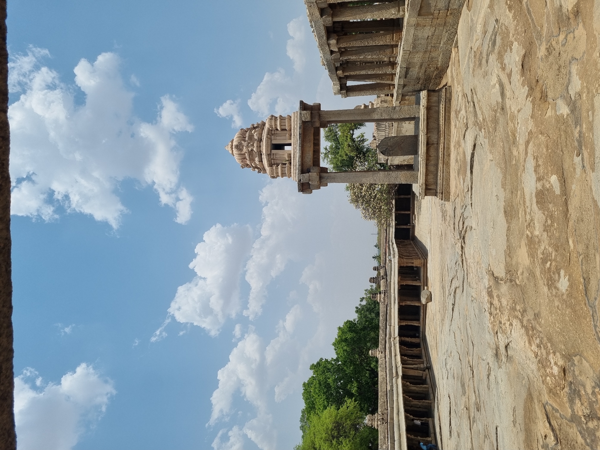 The image depicts a partial view of the Veerabhadra temple in Lepakshi, Andhra Pradesh, India, showcasing its unique architectural features and surrounding environment. In the foreground, a section of the temple's stonework is prominently displayed, exhibiting intricate carvings and detailing. Further back, a tower-like structure stands against a backdrop of a partly cloudy sky, and some trees. The temple's exterior appears weathered, indicating its age and exposure to the elements. The sky is a blue with white clouds, creating a backdrop for the temple structure. The absence of people in the immediate vicinity highlights the architectural and historical context of the temple.