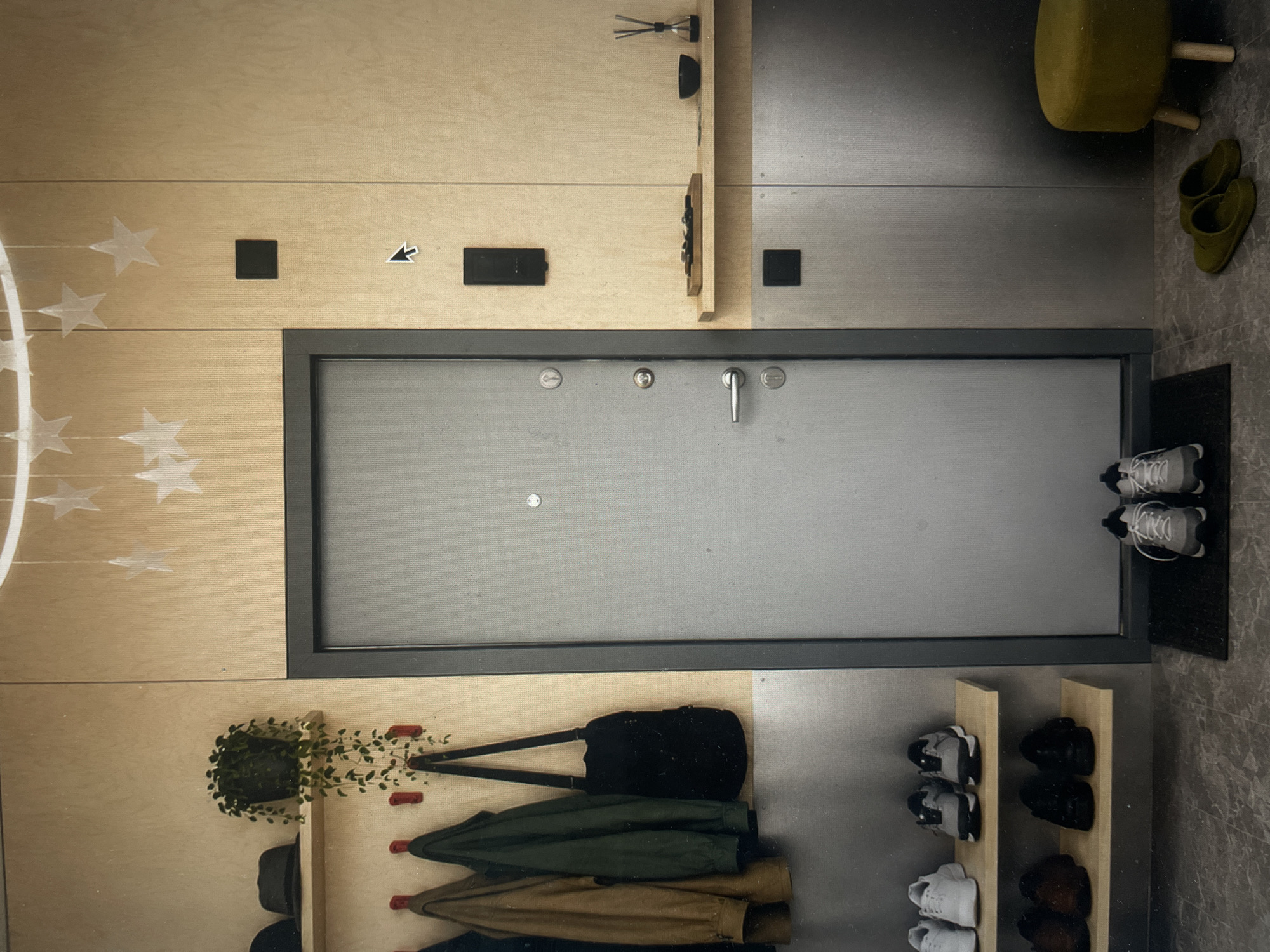 This is an interior shot, likely taken in a residential apartment in Saint Petersburg, Russia, focusing on the entrance area. The primary focal point is the closed metal door with a dark frame, equipped with a silver handle.
Above the door are two small black square objects and a small arrow pointing left affixed to the wooden wall.
A light wood paneling covers a large portion of the wall. On the left side of the wall are decorative star shapes. Below the door is a light wood wall-mounted coat rack, holding several coats in shades of brown and green and a black bag. A green potted plant is visible above the coat rack, next to a black hat.
To the right of the door, the wall transitions to a darker, gray metal. There is a yellow ottoman with wooden legs. On the floor, next to the ottoman, are a pair of green slippers.
Near the bottom right of the frame is a small shoe rack made of light wood. It holds several pairs of shoes in various colors, including black, brown, and gray. A gray doormat sits near the top right, with two pairs of white sneakers placed on it. The floor is tiled with gray marble-like material.