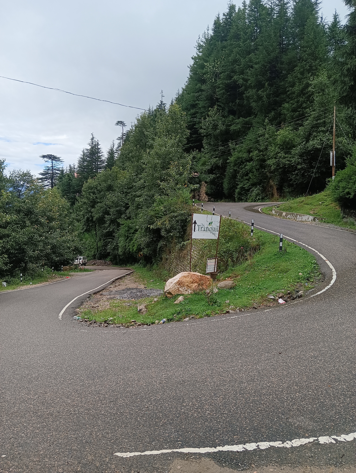 The image shows a winding asphalt road amidst a lush green forest in Goshal, India. The road curves sharply, creating a hairpin turn. On the right side of the road, a signpost indicates "The Tranquil". The area is heavily wooded with tall, dense pine trees. The sky is overcast with a few patches of blue. There are some white road markers and a large rock on the side of the road. A small white vehicle is visible in the distance on the road. The overall scene depicts a scenic mountain road in a natural environment.