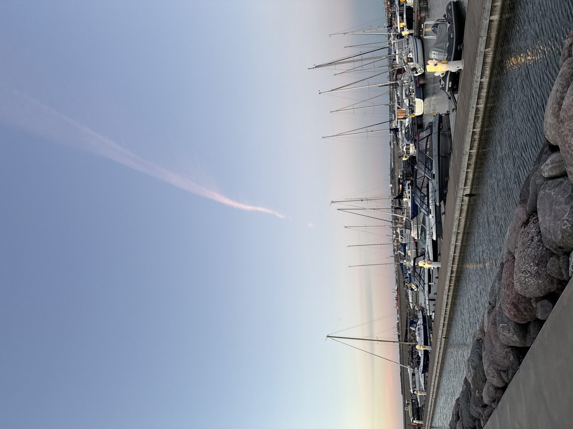 The image depicts a marina in Tallinn, Estonia, likely during sunset or sunrise. Numerous sailboats and motorboats are docked along a wooden pier. The water is calm, reflecting the lights from the boats and the sky. A breakwater made of large rocks lines the foreground, separating the marina from a concrete walkway. In the sky, a faint contrail stretches across the pale blue expanse, tinged with pink and orange at the horizon. The overall scene is peaceful and serene, showcasing a typical evening at a coastal city harbor.