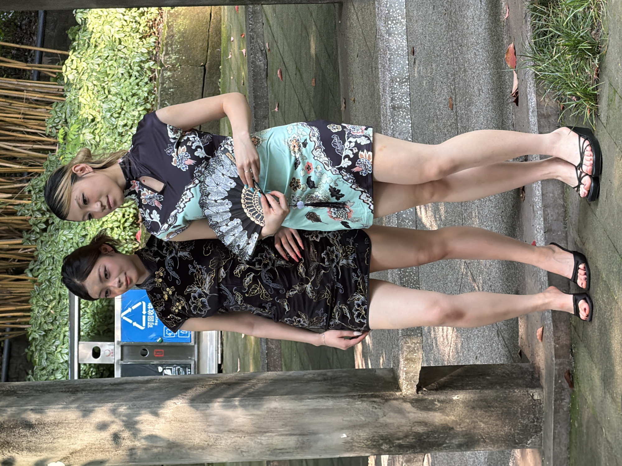 Two young East Asian females are standing outdoors in Yuzhong District, China, in what appears to be a park or garden setting. Both are wearing short-sleeved, high-collared mini-qipaos and are positioned as if posing for a photograph.
The woman on the left wears a black qipao with gold and silver floral and leaf patterns. She has dark hair styled back and looks slightly to her right. Her right hand, with bright orange nail polish, is placed on her hip. She wears a silver bracelet on her left wrist and black open-toe mules with red nail polish on her toes.
The woman on the right wears a two-toned qipao, featuring a dark blue upper section and an aqua lower section, both adorned with light-colored floral and wave patterns. Her dark hair with prominent blonde highlights is pulled back, and she looks slightly to her left. She holds an open black folding fan with intricate white and light blue floral designs in her hands, from which a dark tassel hangs. She wears black wedge-heeled flip-flops with multiple thin straps and red nail polish on her toes.
They are standing on a textured concrete surface with visible patches of moss and scattered fallen leaves. Behind them, to the left, is a large, grey concrete pillar. Further back, on the left, are two public waste receptacles; one is dark grey, and the other is blue with a white triangular recycling symbol and the Chinese characters "回收" (huíshōu), meaning "recycle." Behind the receptacles and extending across the midground is a dense layer of green foliage, likely on a raised planter or wall. In the background, above the foliage, numerous slender, vertical brown bamboo stalks are visible. The scene is lit by natural daylight, casting shadows.