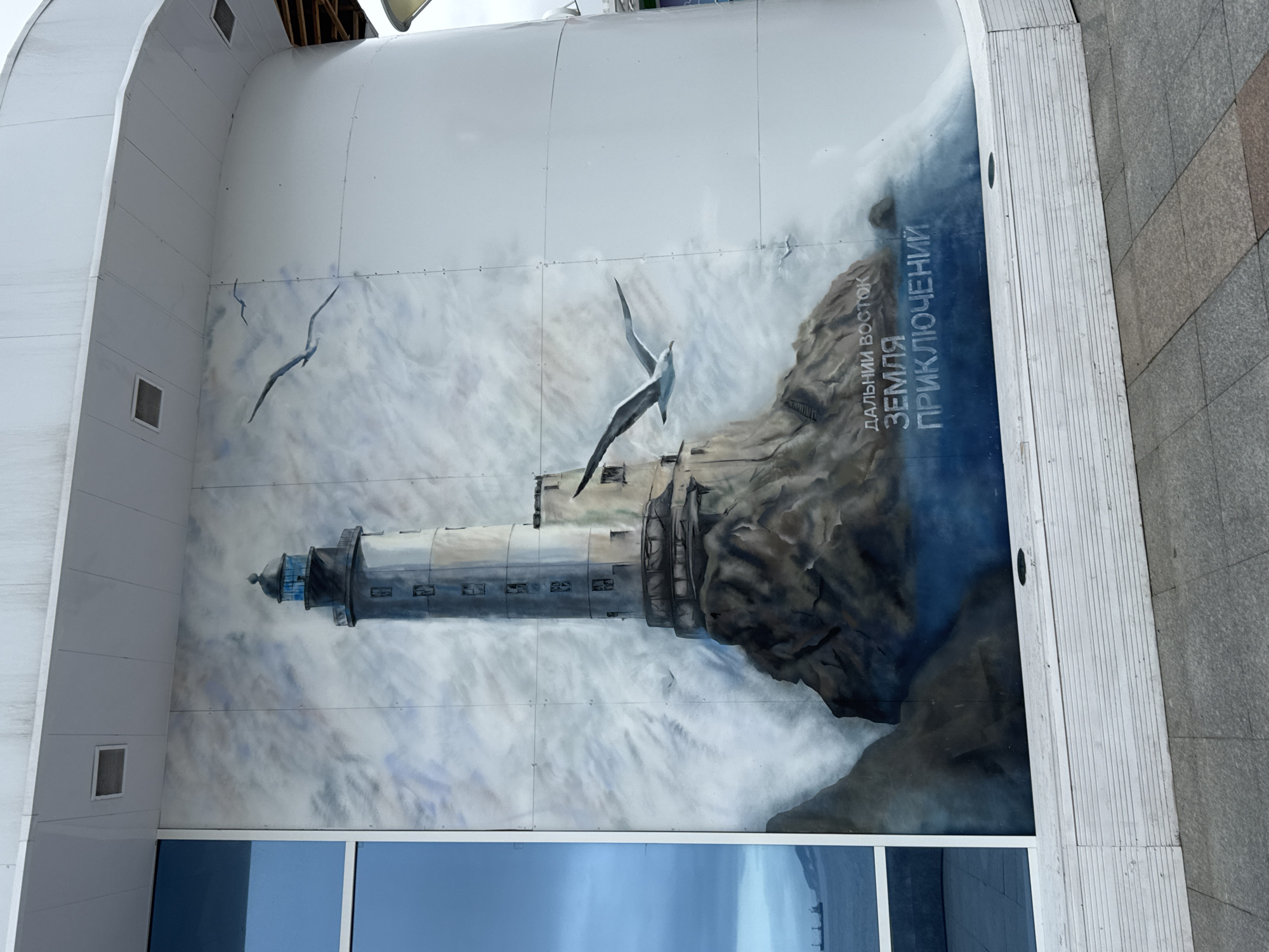 The image displays a mural painted on the exterior curved wall of a building, situated in the city of Russky, Russia. The mural depicts a lighthouse situated on a rocky outcrop, with waves crashing around its base. Two seagulls are painted in flight near the lighthouse. The background consists of a cloudy sky rendered in shades of blue and white. Below the mural, text is visible in Cyrillic letters. To the left of the mural is a large, reflective blue window. The building appears to be modern, with a white facade. The ground in front of the building is paved with grey and brown tiles. The scene is captured from a slightly elevated perspective, looking up at the mural.