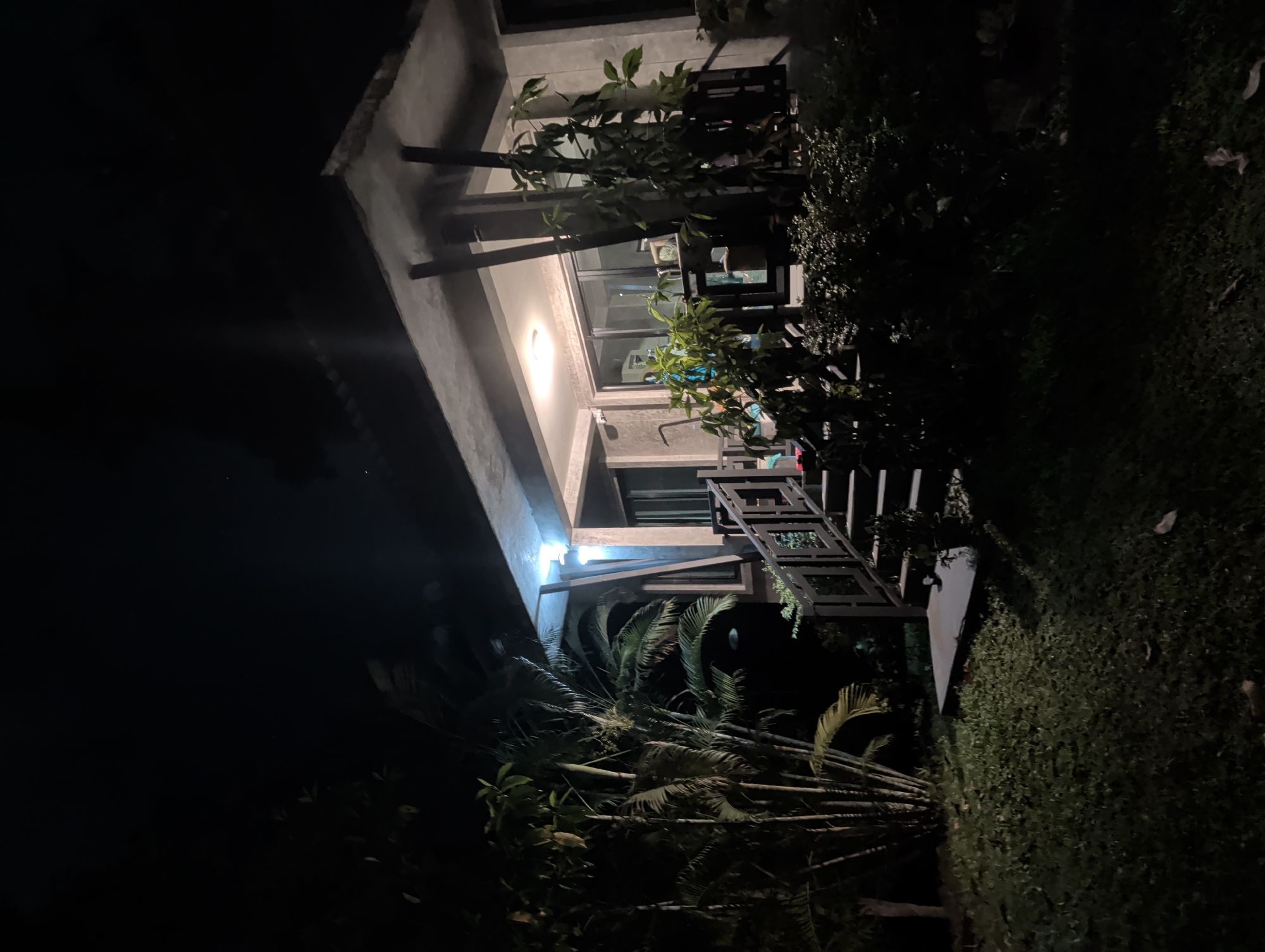 The image shows a building at night in ตำบลเกาะพะงัน, Thailand. The building is multi-story with a concrete structure. The lighting is dim, primarily from interior lights and external fixtures. A staircase with metal railings is visible, connecting different levels. Vegetation, including trees and shrubs, surrounds the building. The scene appears to be a residential or resort area with landscaping. The image is taken from a low angle, looking up at the building.