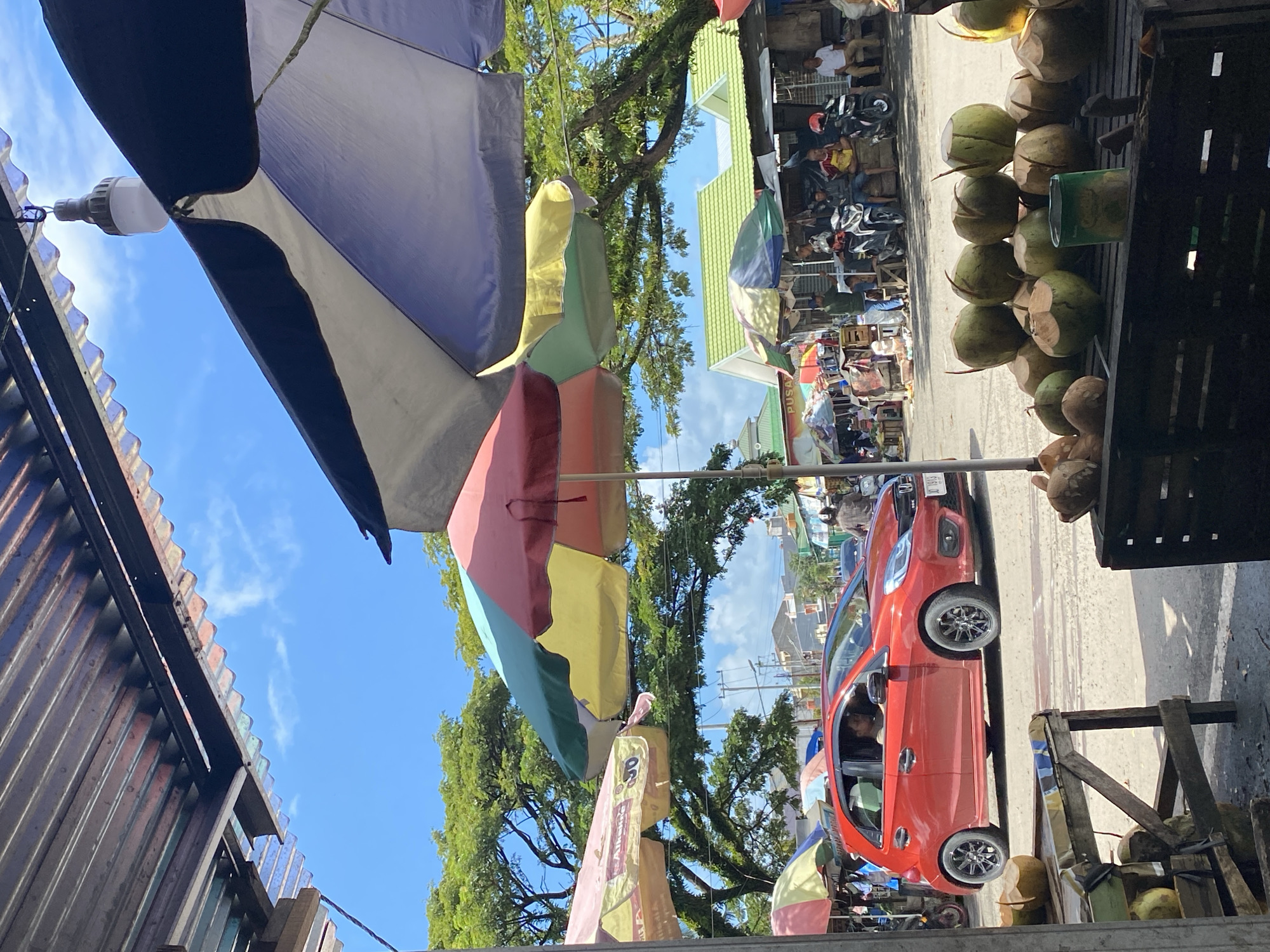 This image depicts a vibrant street scene in Ta', Indonesia, likely a local market on a sunny day. The foreground is dominated by a display of fresh coconuts arranged on wooden crates. To the left, a portion of a wooden stall structure is visible.
In the midground, a red hatchback car is parked, with a person visible in the driver's seat. Behind the car, a street stretches out, lined with various market stalls covered by colorful umbrellas and tarps. People are milling about, some sitting, some standing, and a few motorcycles are parked. The stalls appear to be selling goods, as indicated by the merchandise and the presence of customers.
The background features more buildings, some with green roofs, and lush green trees that arch over the street, providing shade. The sky is bright blue with scattered white clouds, suggesting a clear and pleasant weather day. The overall impression is a bustling, open-air marketplace, characteristic of many towns in Indonesia.